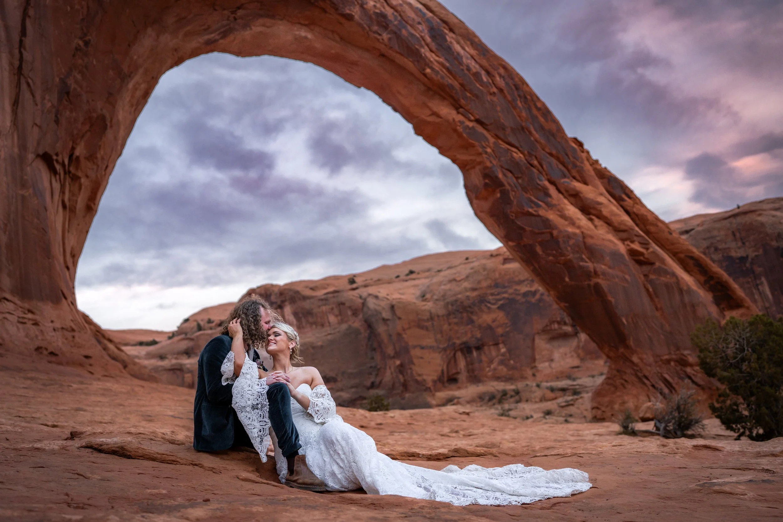 A couple in wedding attire sitting on the desert ground beneath a large natural rock arch.