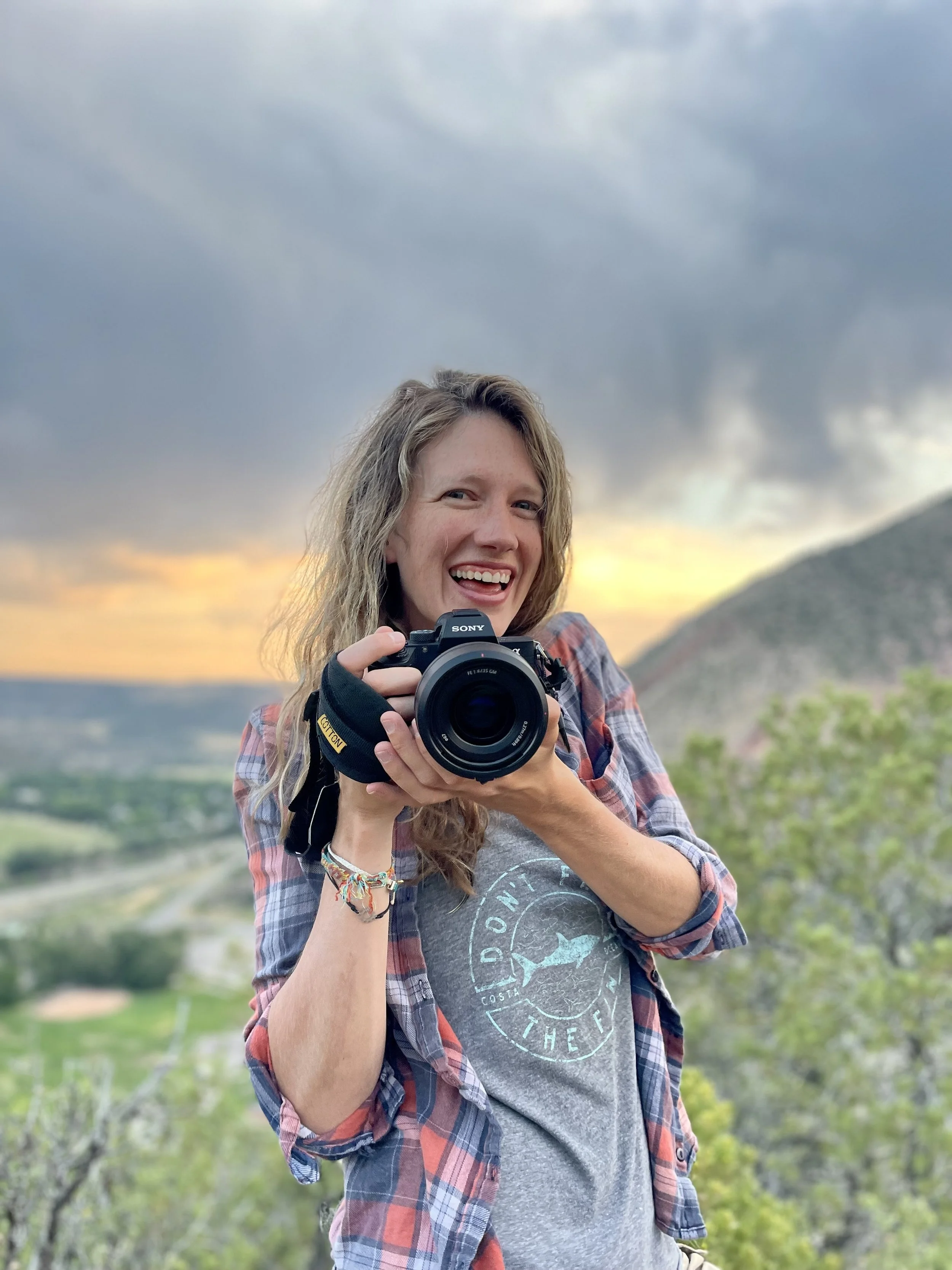 A woman smiling and holding a camera outdoors at sunset with hills and trees in the background.