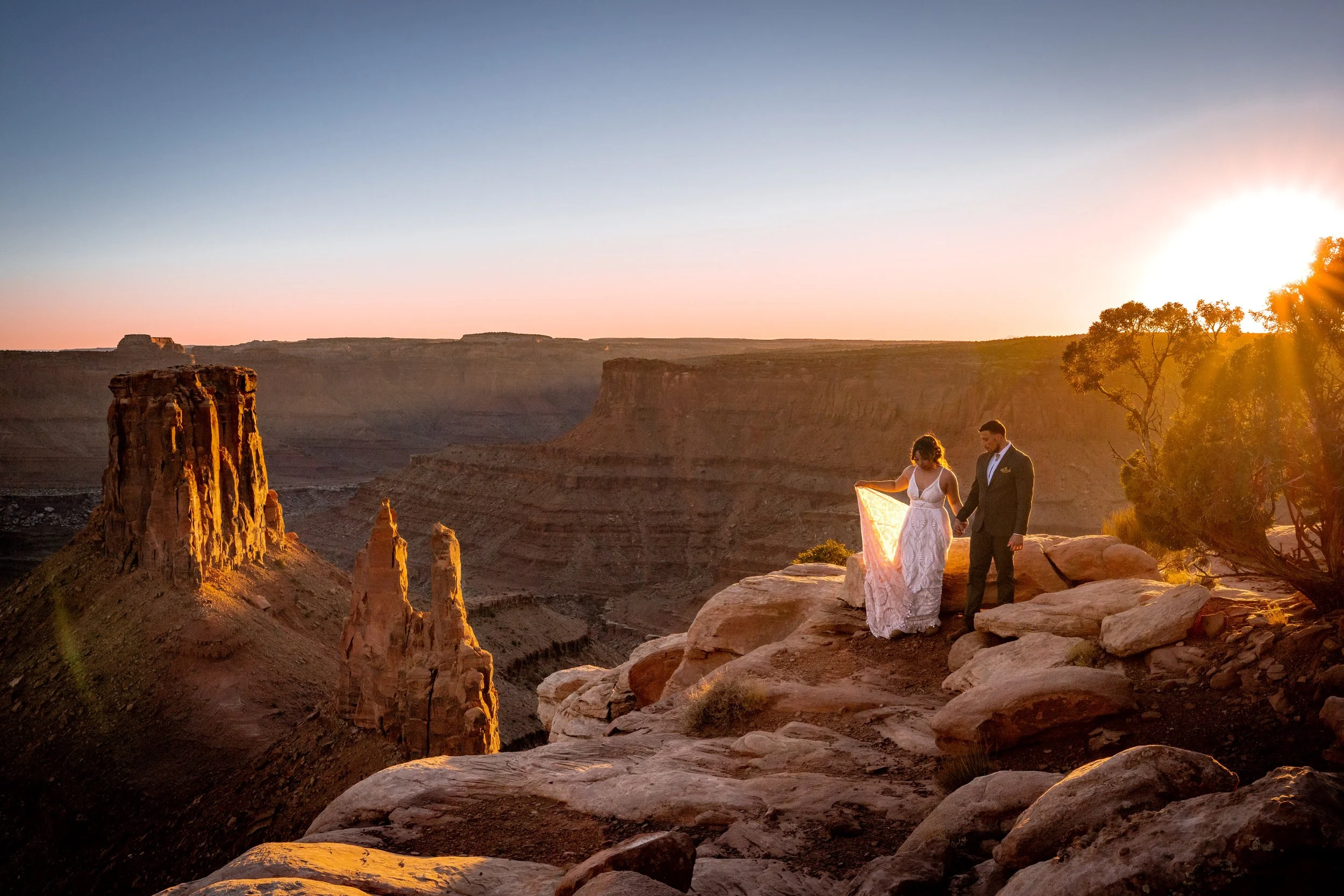 A bride and groom walk together along a red rock Utah canyon at sunset.