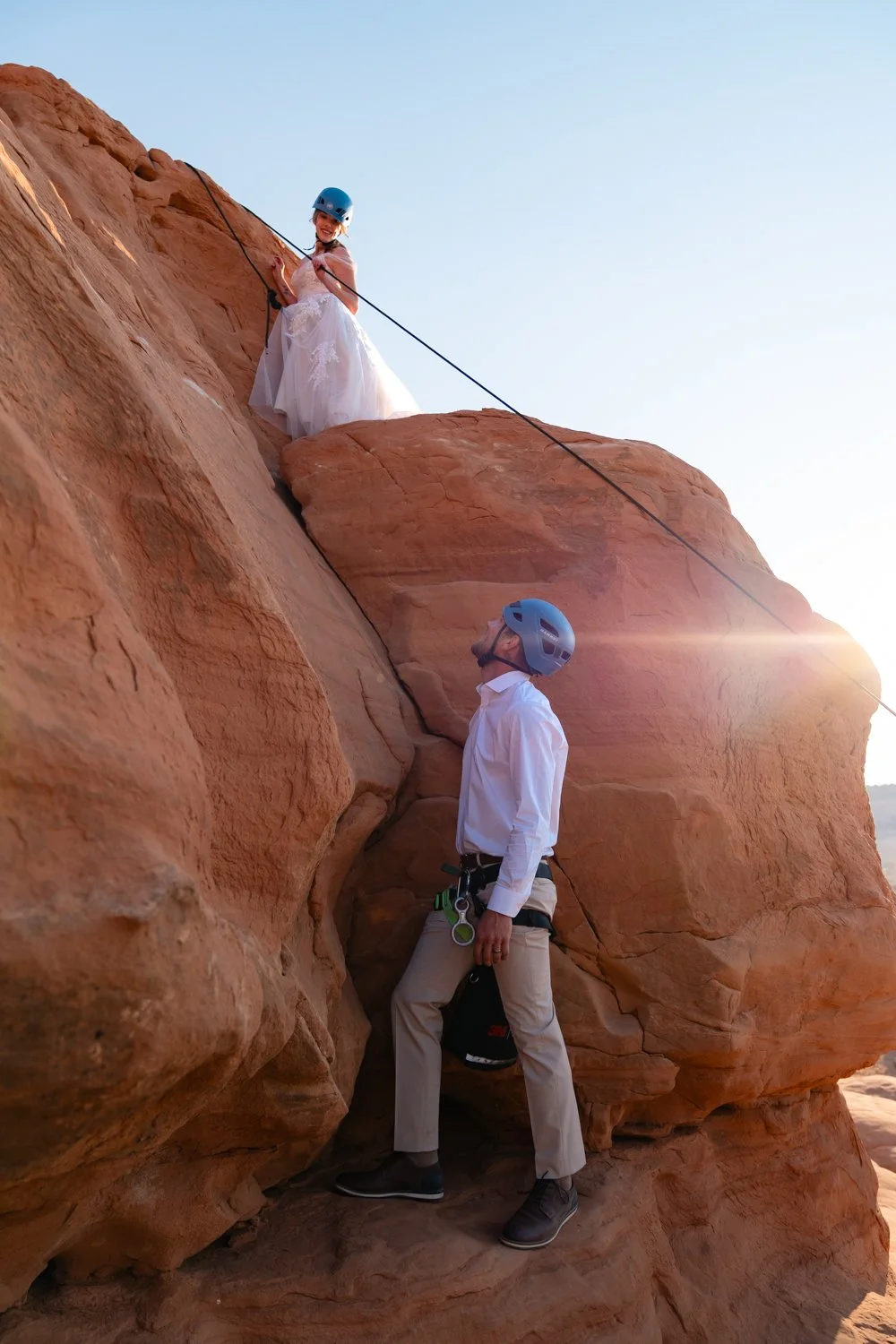A bride looks down at a groom from a rock as they climb together in Moab.