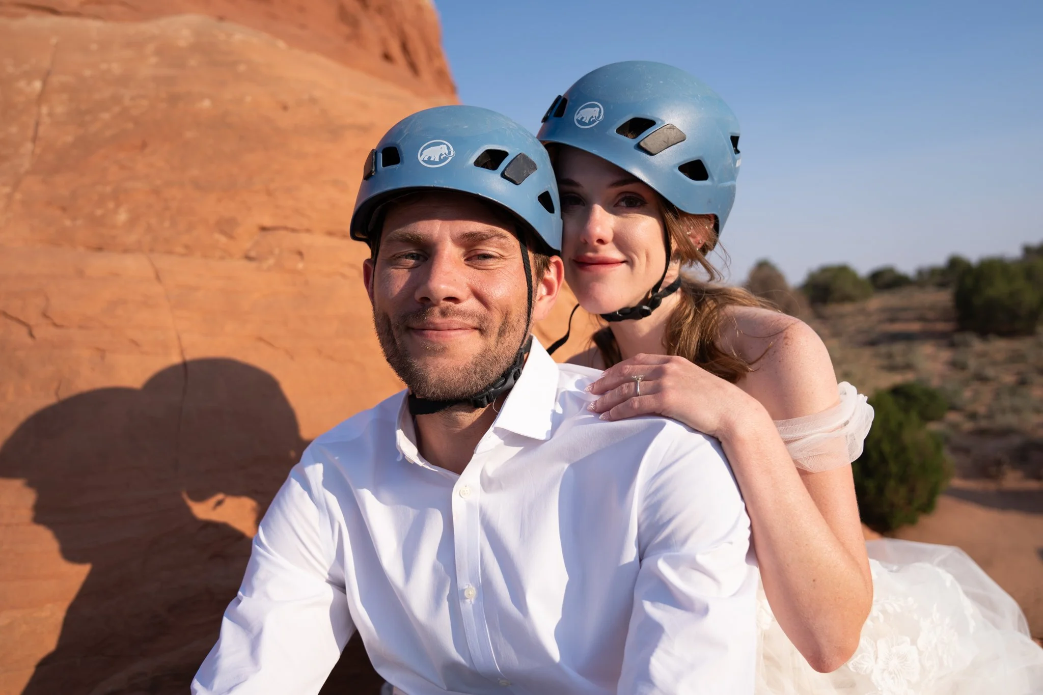 A bride and groom smile at the camera in climbing helmets.