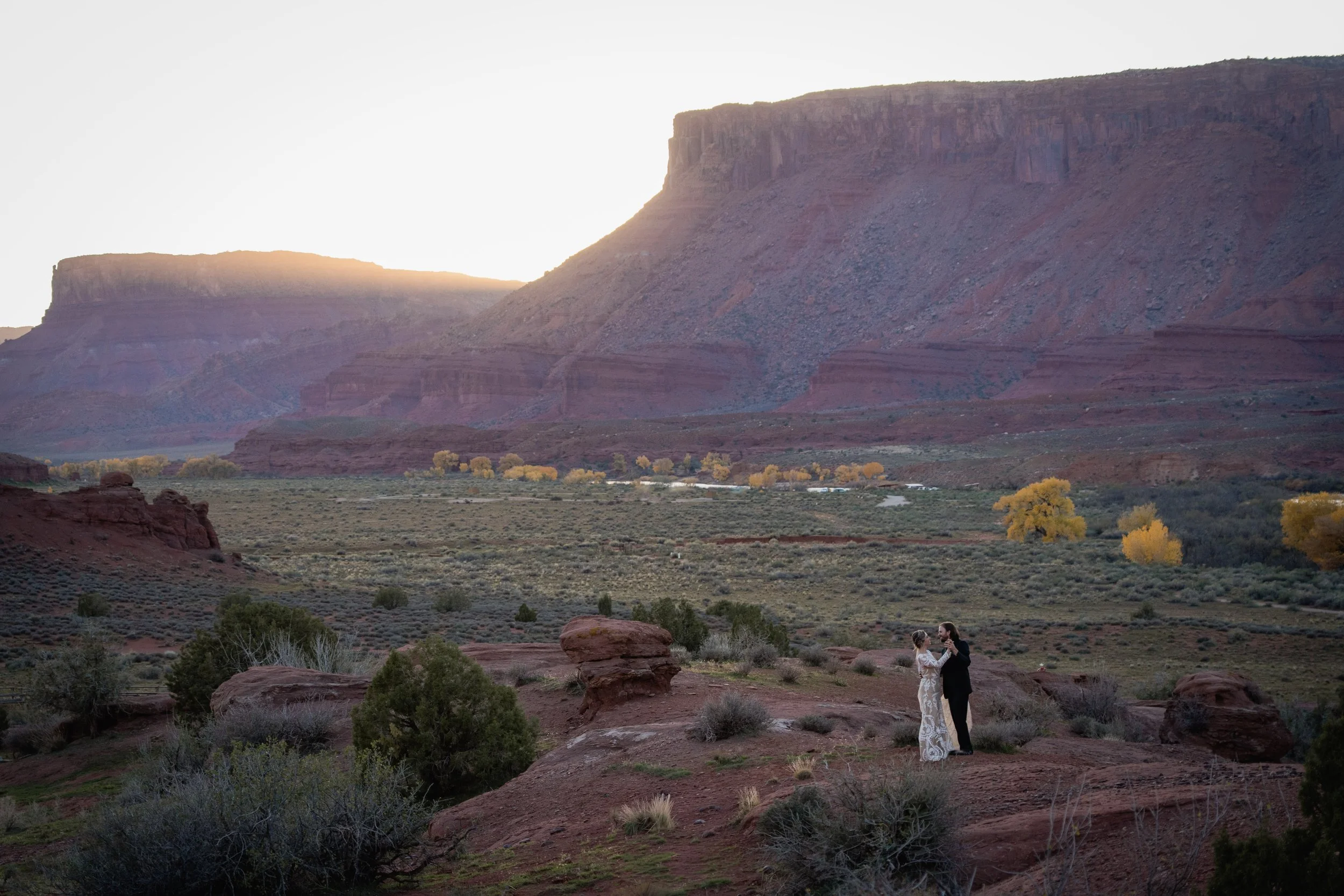 A bride and groom dance in the desert under canyon walls at sunset.