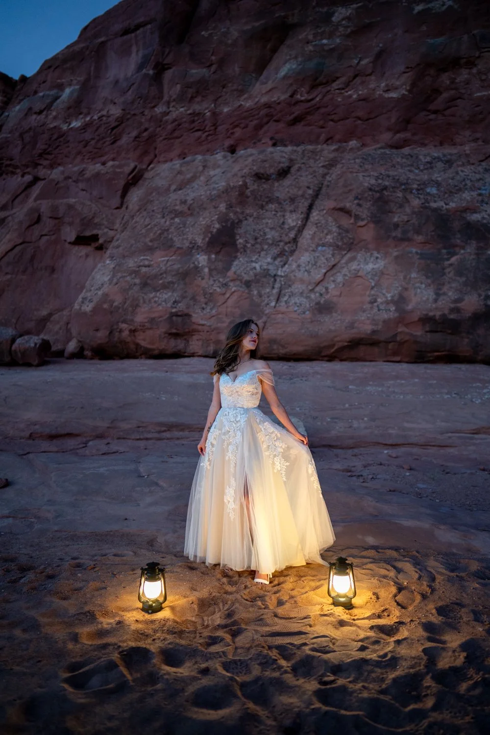 A bride looks off into the distance as she holds her wedding dress against lantern light and red rocks in Moab.