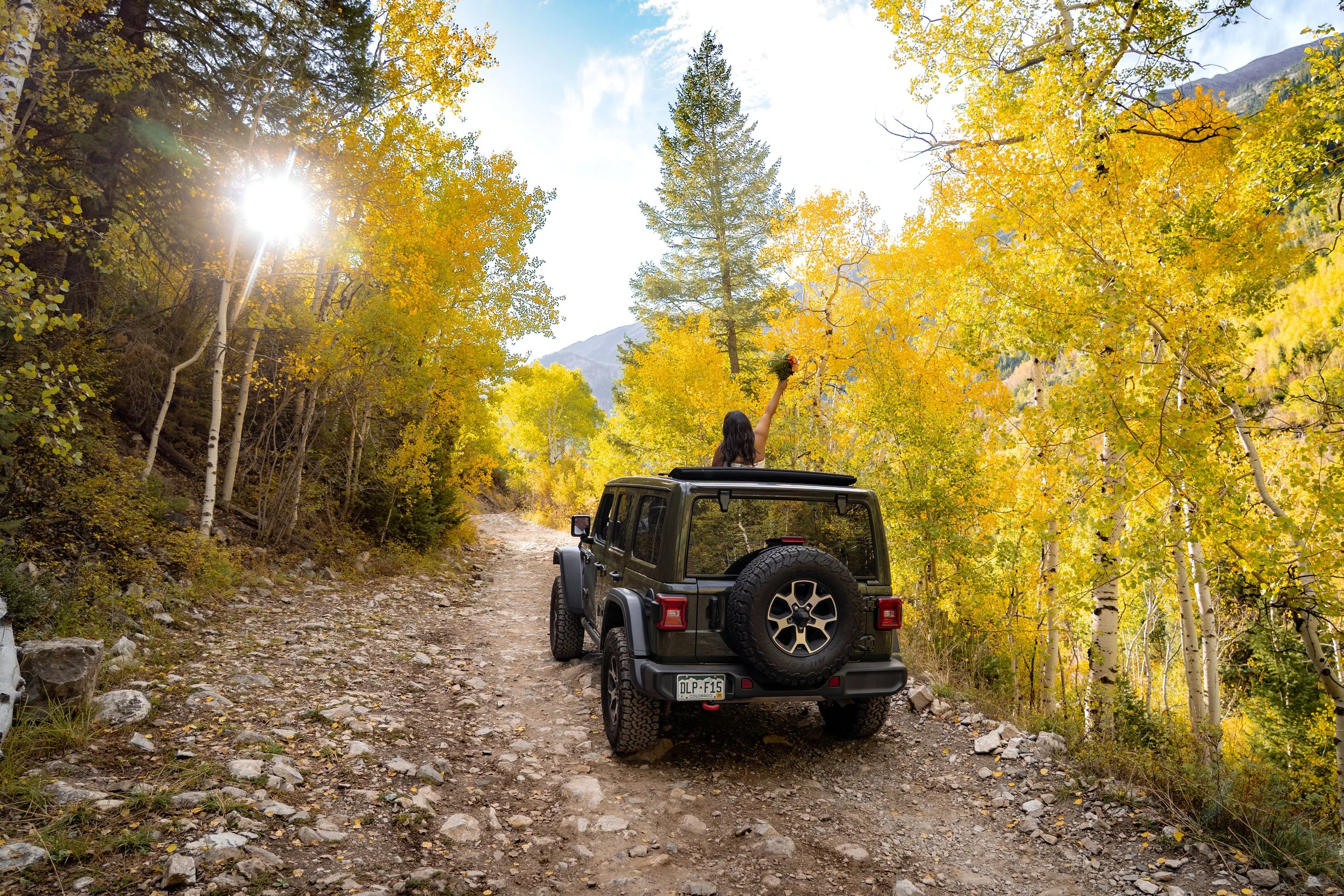 A bride holds her flower bouquet outside of the top of a Jeep while offroading down a trail in fall.