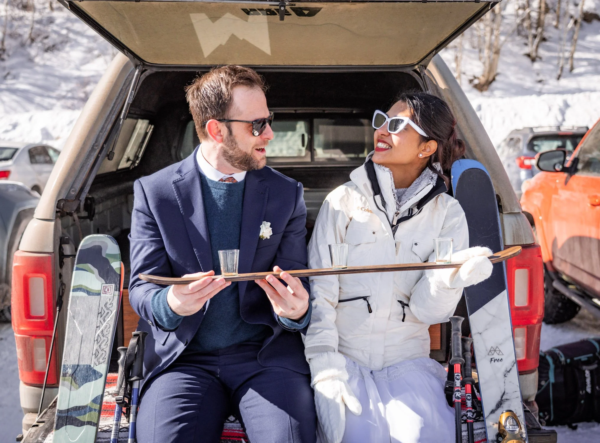 A bride and groom smile at each other from the back of a car at a ski resort.