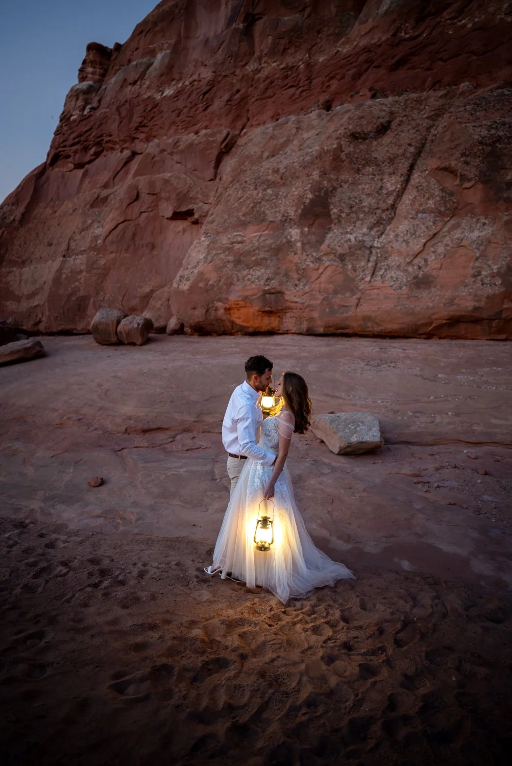 A bride and groom stand close with lanterns in front of red rocks in Moab, Utah.
