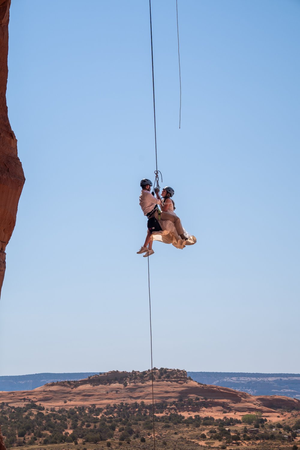A bride and groom rappel down together laughing from a rock in Moab, Utah.