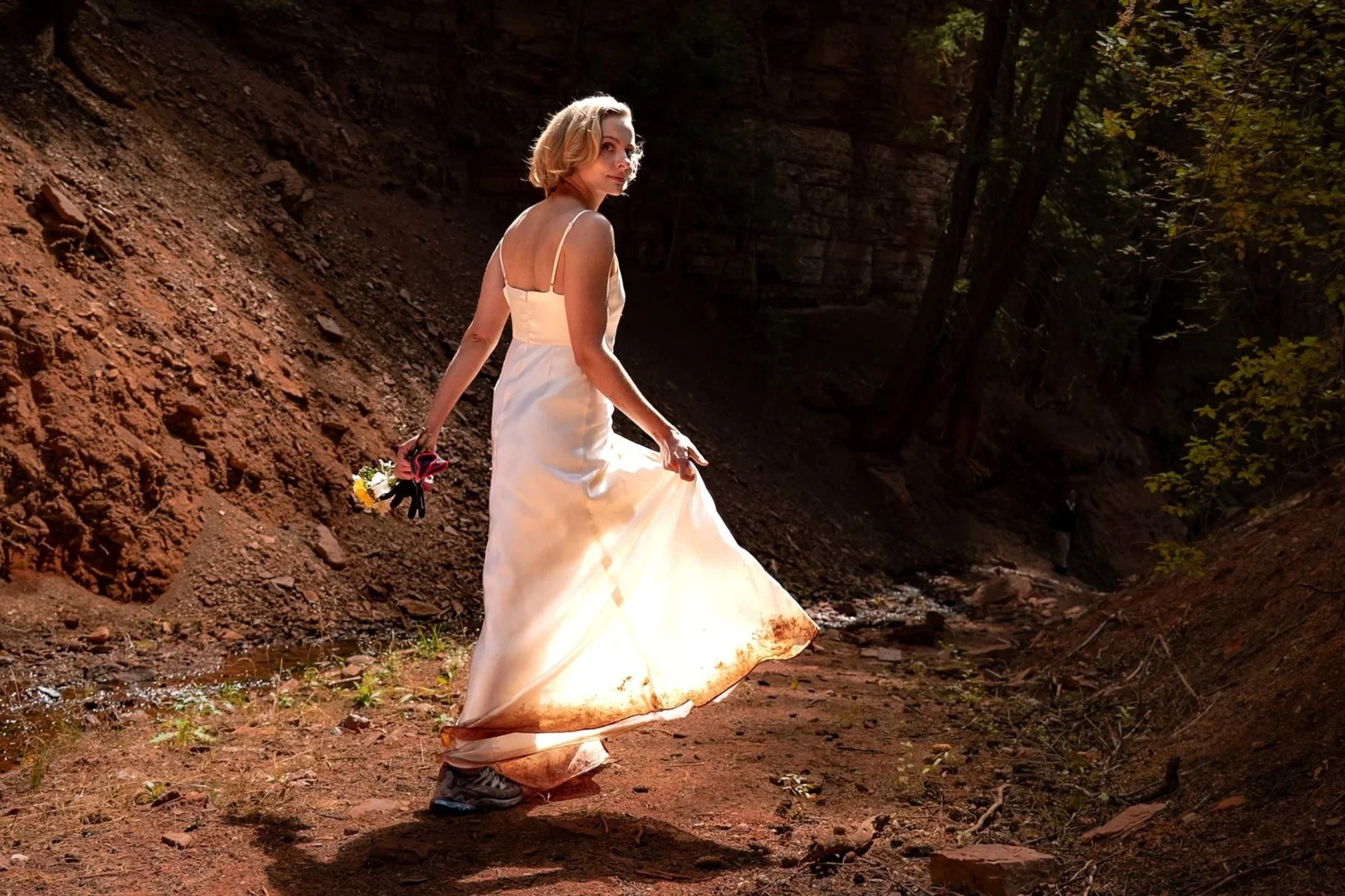 A bride holds out her sunlit wedding dress while revealing hiking boots.