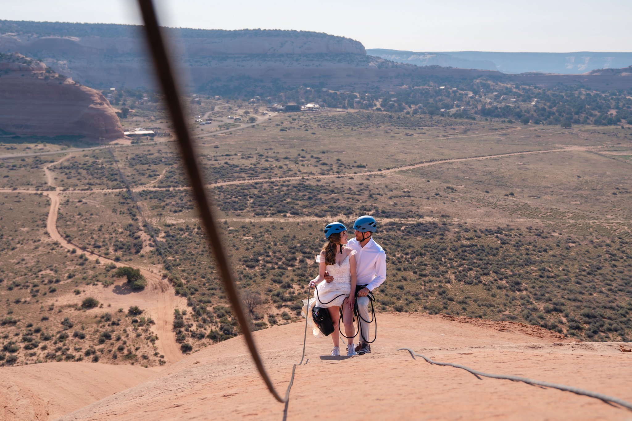 A bride and groom stand close looking at each other as they climb up a rock in Moab.