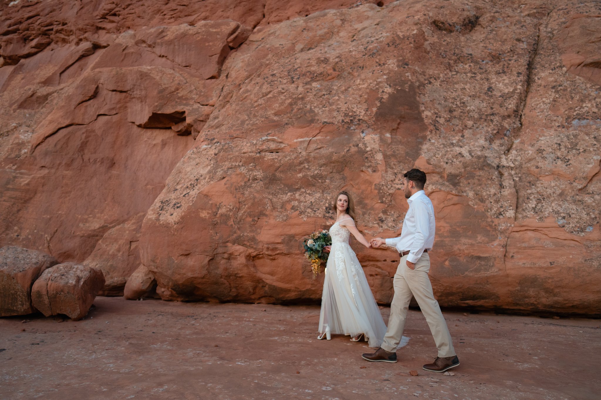 A bride leads a groom walking over red rock in Moab.