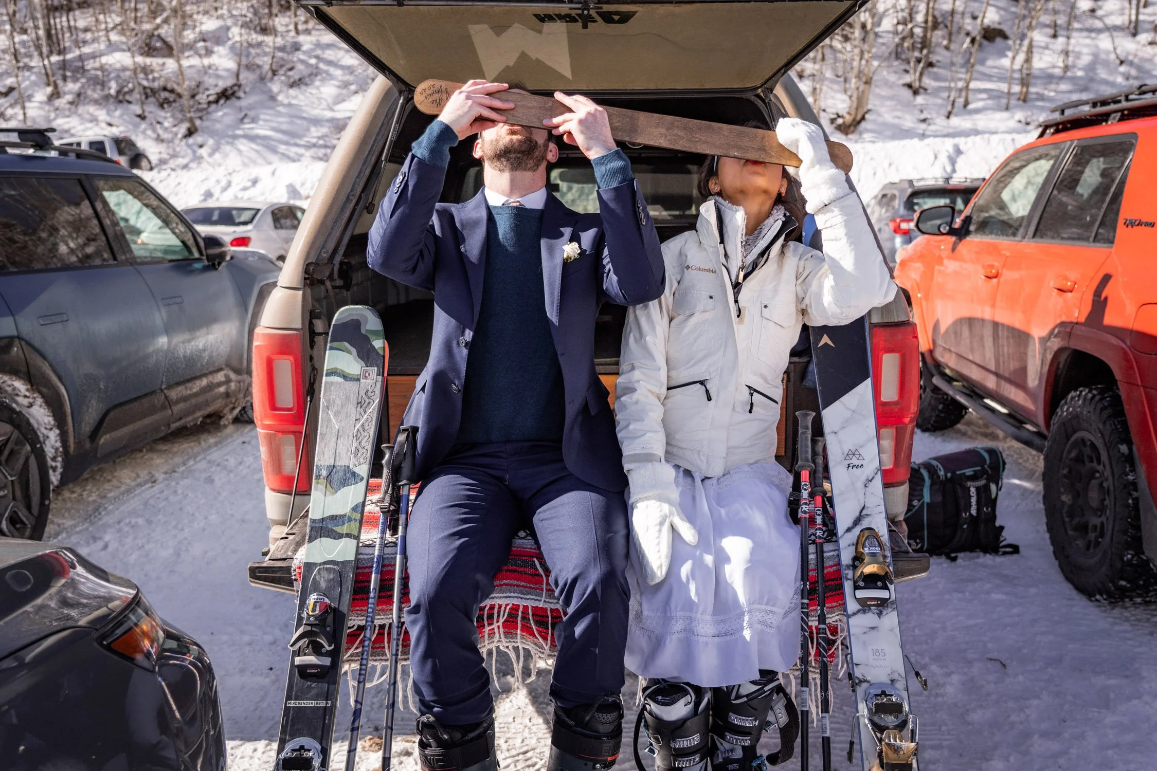 A bride and groom take a shot ski on the back of a car at a ski resort.