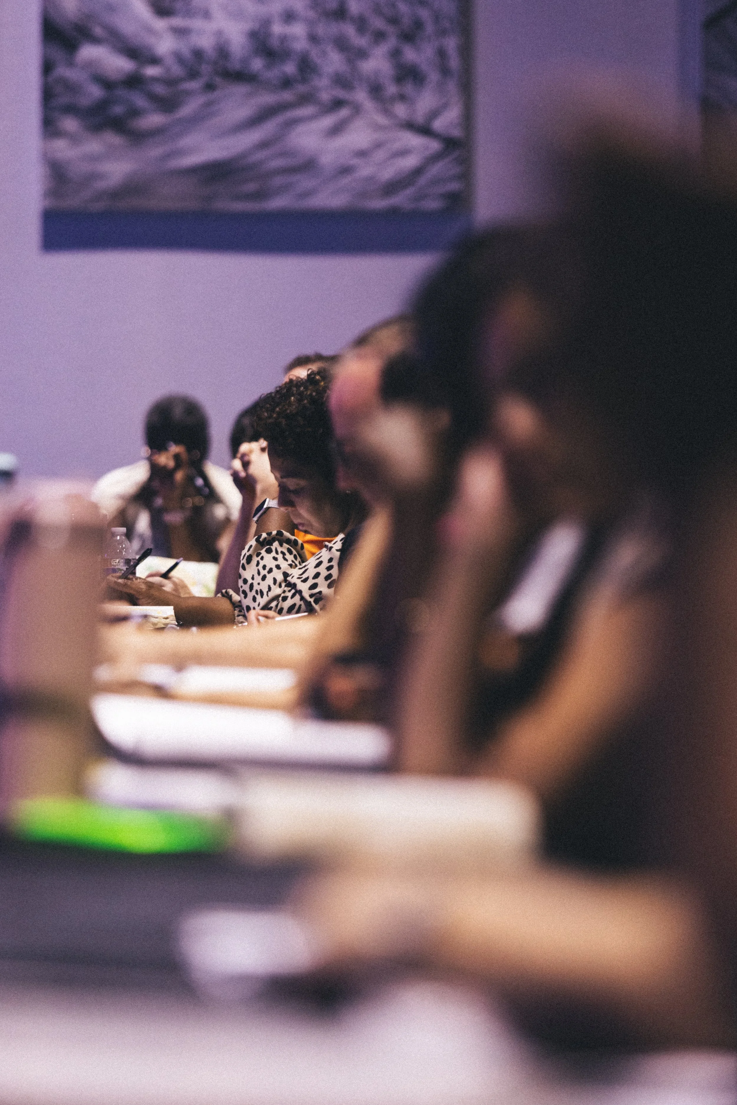 Several people sitting at a table, appearing to be attending a conference or seminar, with some taking notes and others looking at their phones, in a dimly lit room with purple walls and artwork.