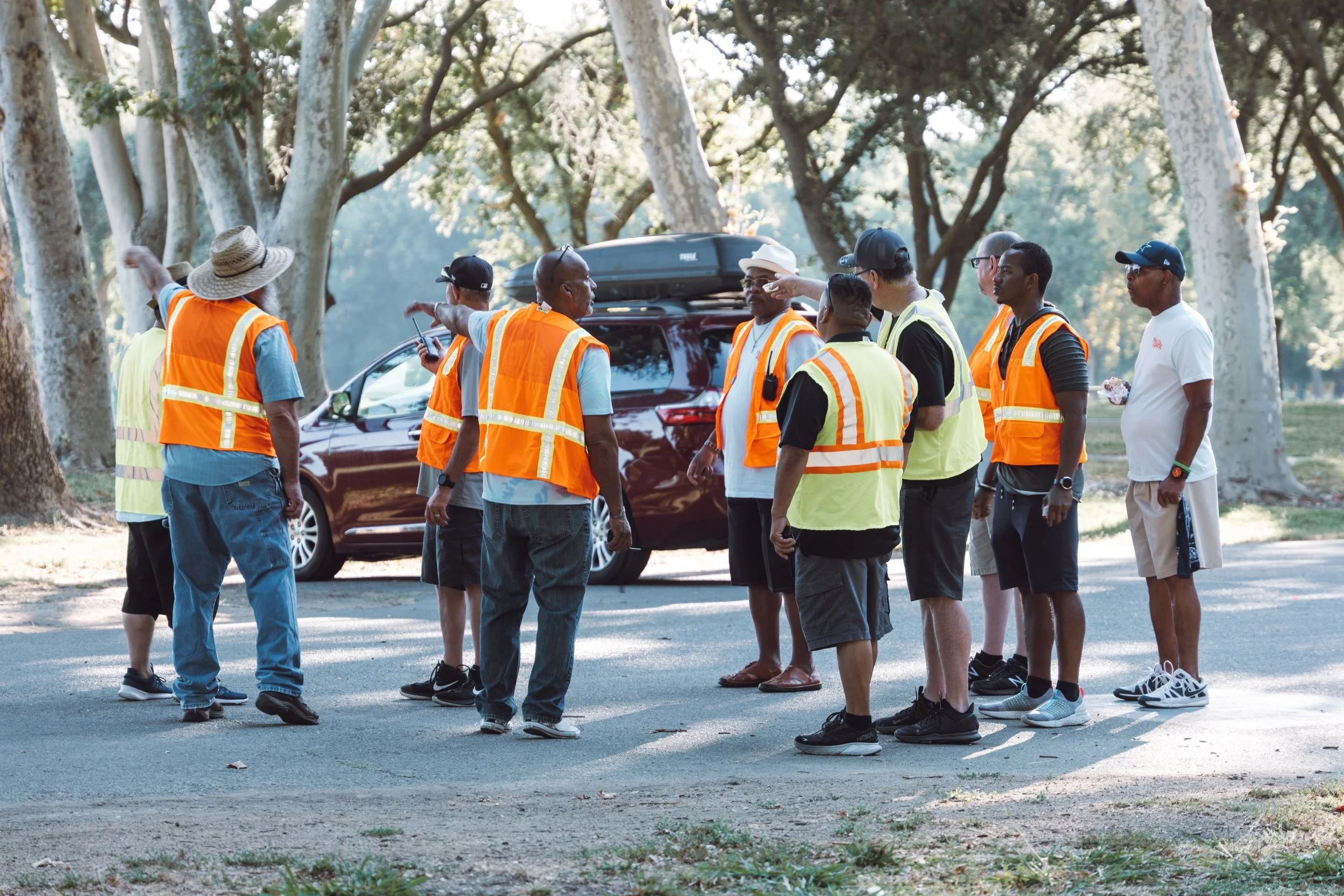 Group of people, some wearing orange safety vests, gathered around in a park with large trees. They appear to be having a discussion near a dark-colored vehicle.
