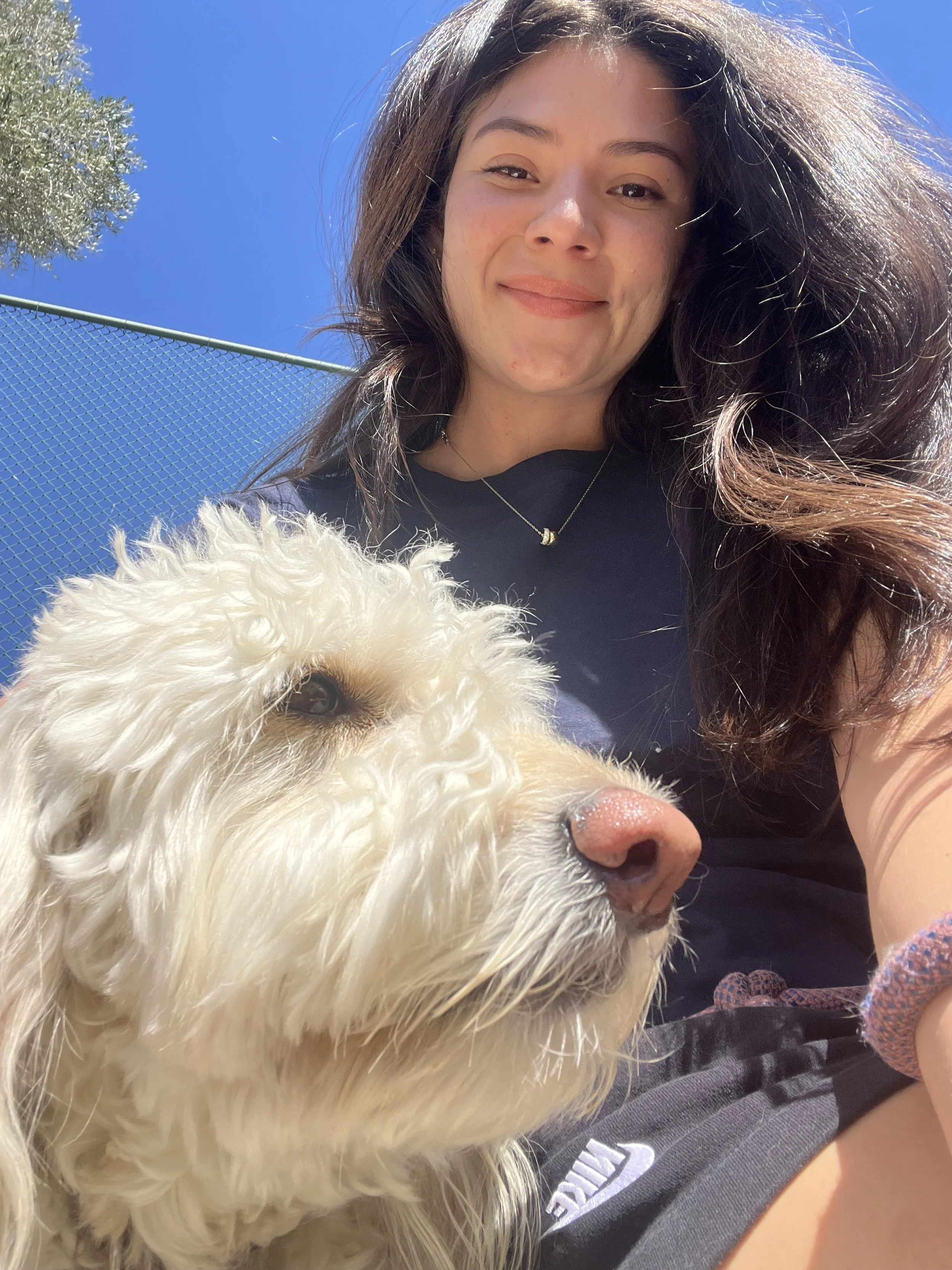 A young woman with long, wavy brown hair takes a selfie outdoors on a sunny day with her fluffy white dog. The woman is smiling, wearing a dark shirt and a necklace, with a bright blue sky and a chain-link fence in the background.