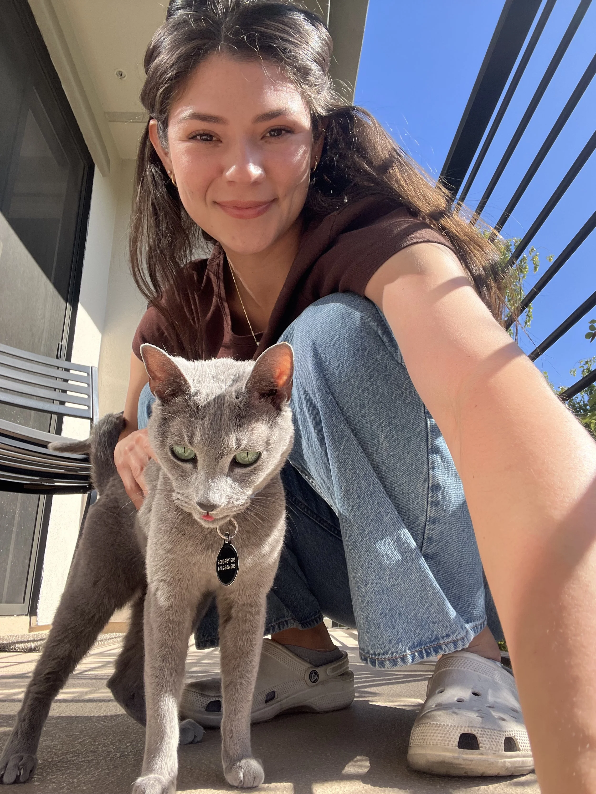 A woman with dark hair and a brown shirt crouches outside on a balcony with clear blue sky, smiling at the camera, next to a gray cat with green eyes and a black collar.