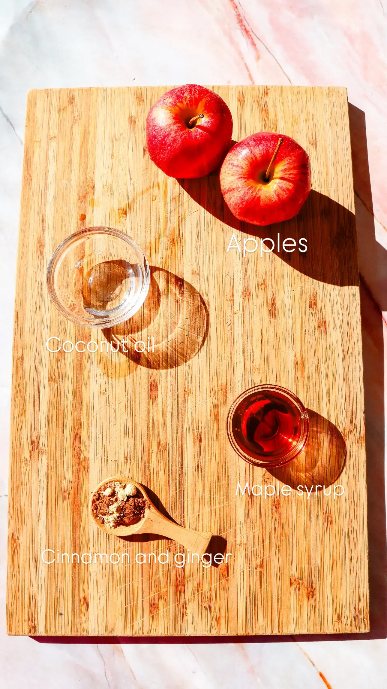 Ingredients for air fryer apples on a cutting board, including apples, coconut oil, maple syrup, cinnamon, and ginger.