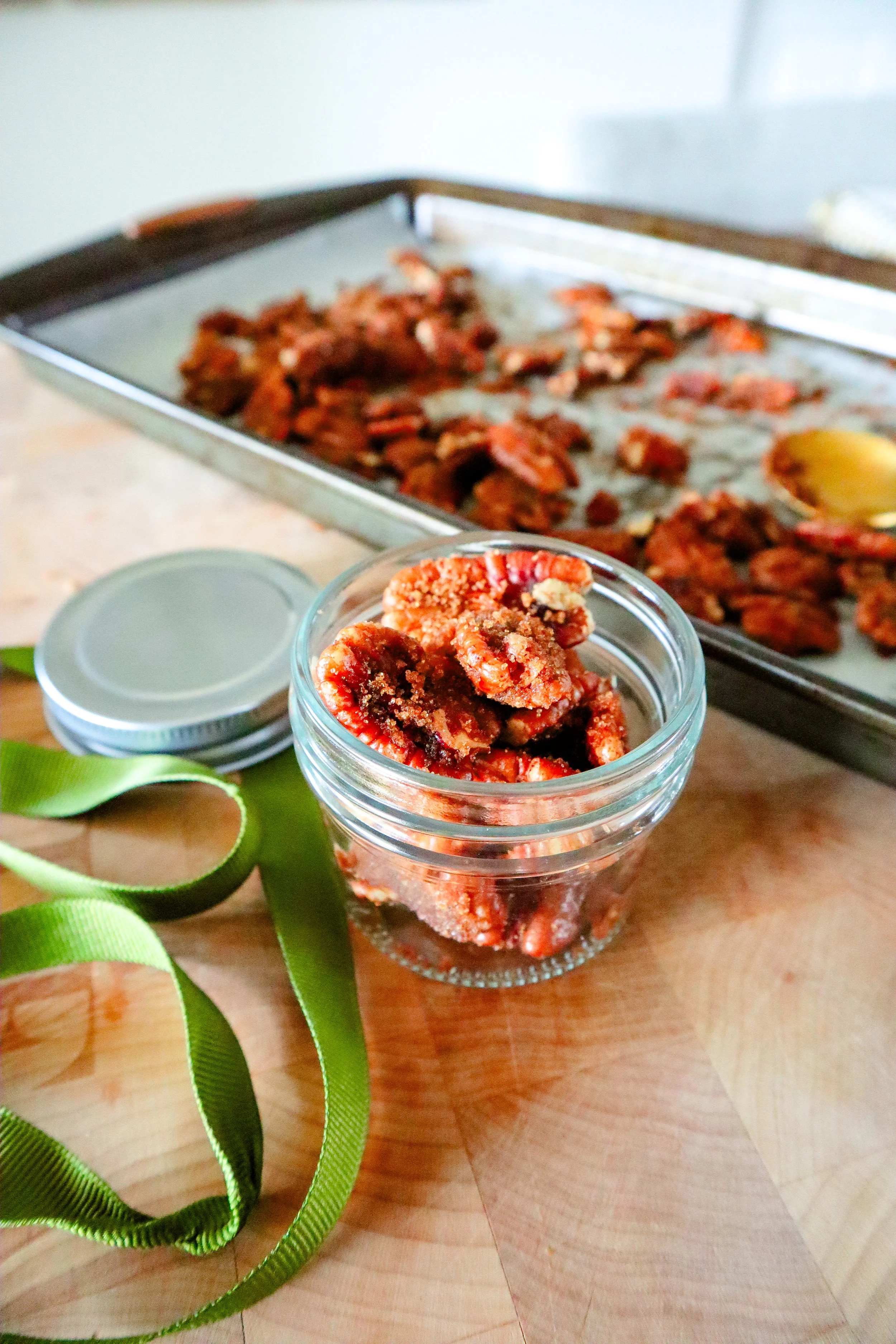  Small glass jar filled with crunchy candied pecans next to a tray of cooled nuts. 