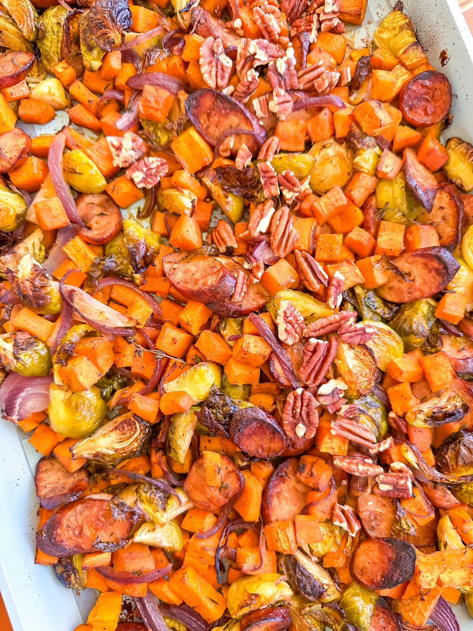  Close-up of a sheet pan filled with roasted sweet potatoes, Brussels sprouts, red onions, and pecans, all lightly browned. 