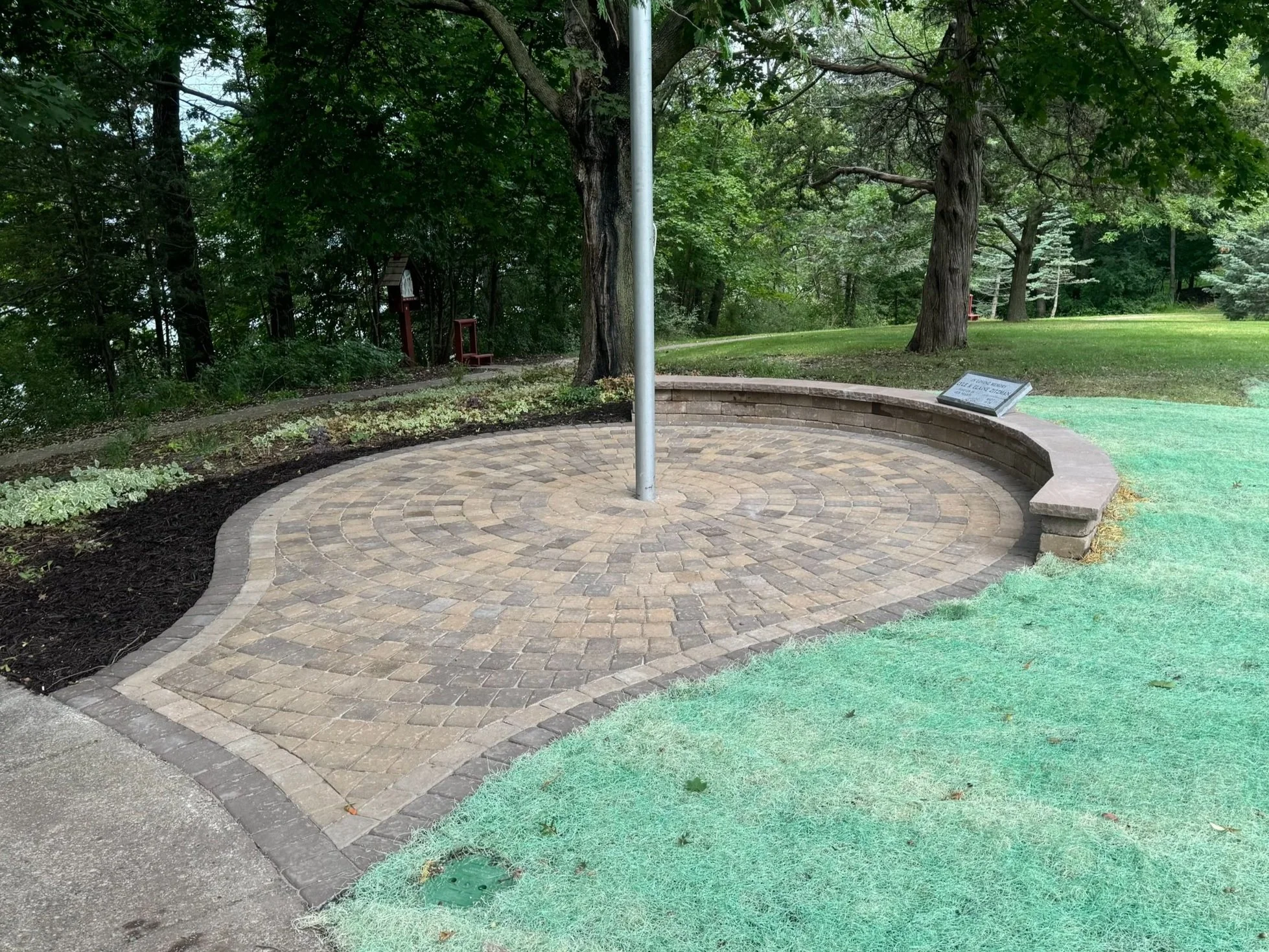 MB Landscapes installed new pavers with a flagpole, circular brick paving, a curved stone bench, and an information plaque in a park surrounded by trees and greenery.