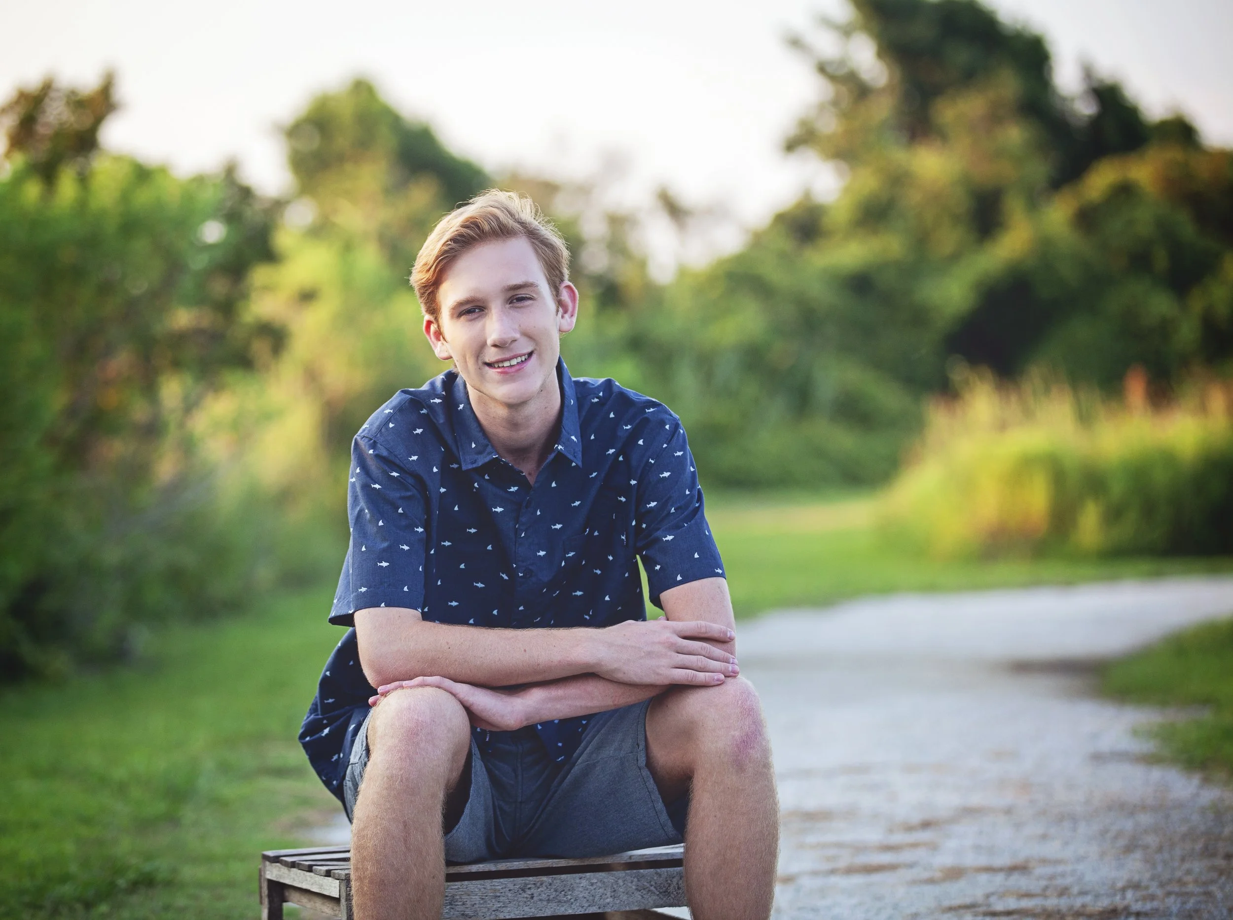 A young man with light brown hair, wearing a navy blue shirt with small white shark patterns and gray shorts, sitting on a wooden bench on a gravel path in a park or natural setting with trees and greenery in the background, during daytime.