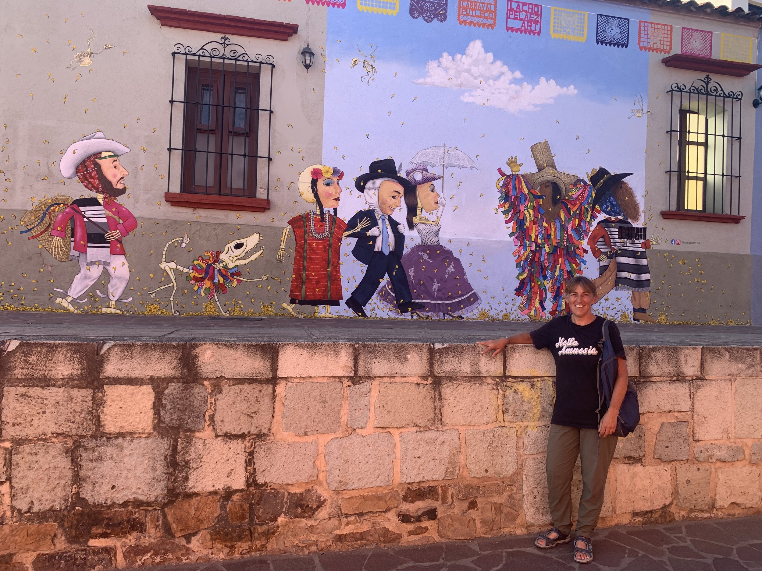  Carla is standing in front of a brightly painted mural in Mexico that shows a parade of skeletons wearing fancy clothing and masks. She is wearing a black t-shirt with ‘Hello Amnesia’ written on it and khaki pants and is smiling.  