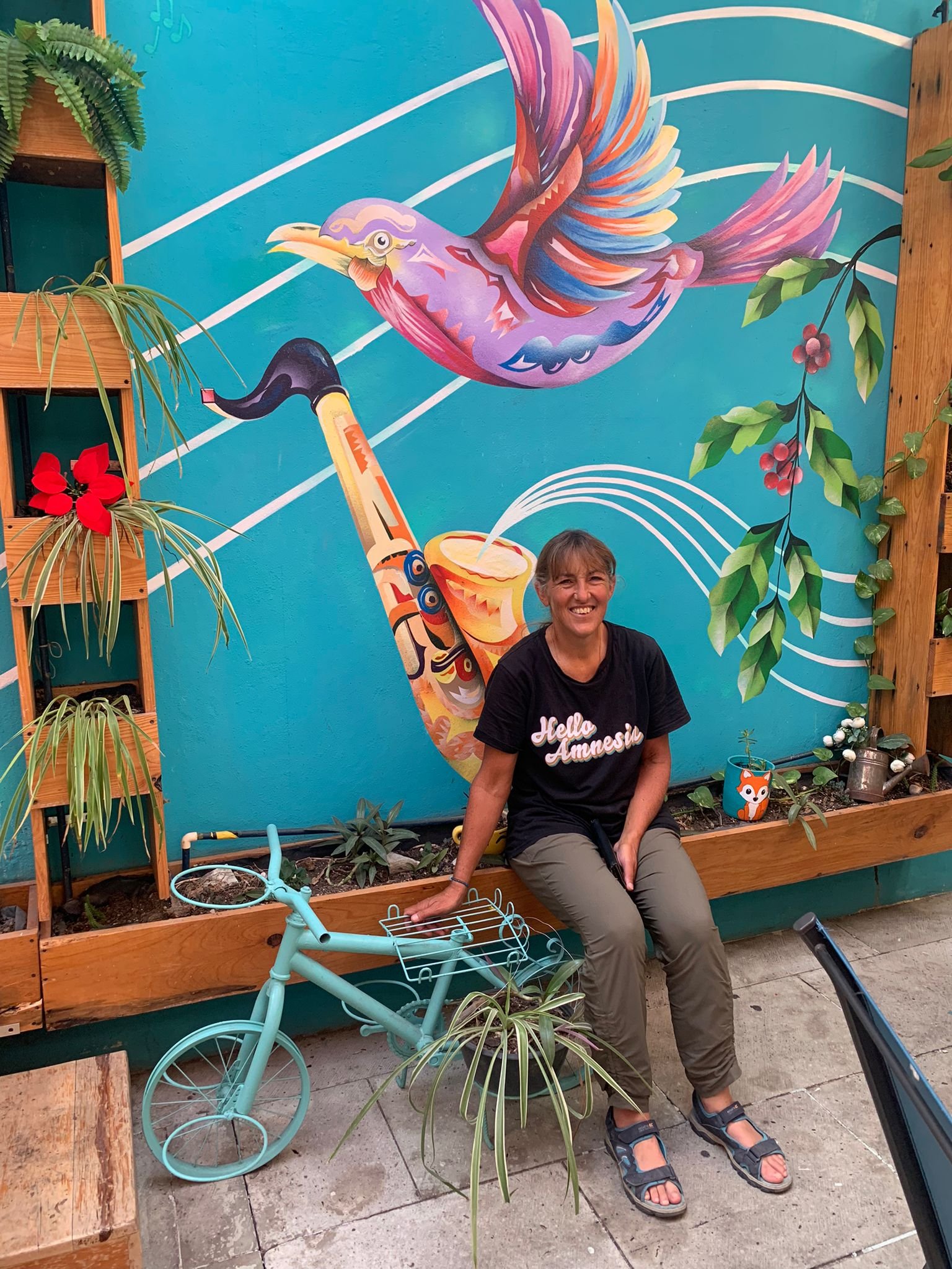  Carla is sitting in front of a mural in Mexico. It has a bright blue background with a multi-coloured bird and a saxophone painted on it. Next to her is an ornamental planter shaped like a bicycle. She is wearing a black t-shirt with ‘Hello Amnesia’
