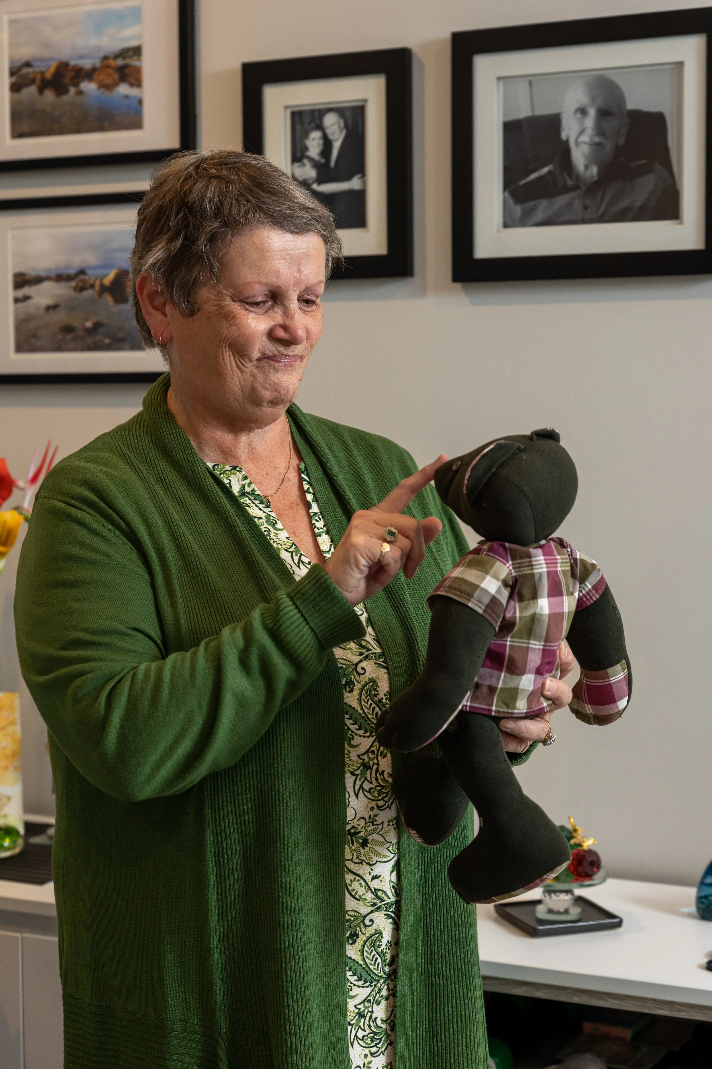 A woman in her 60s wearing an olive green cardigan holding a black teddy bear that’s wearing a plaid shirt. She has her finger on the teddy’s nose and she looks sad and thoughtful.