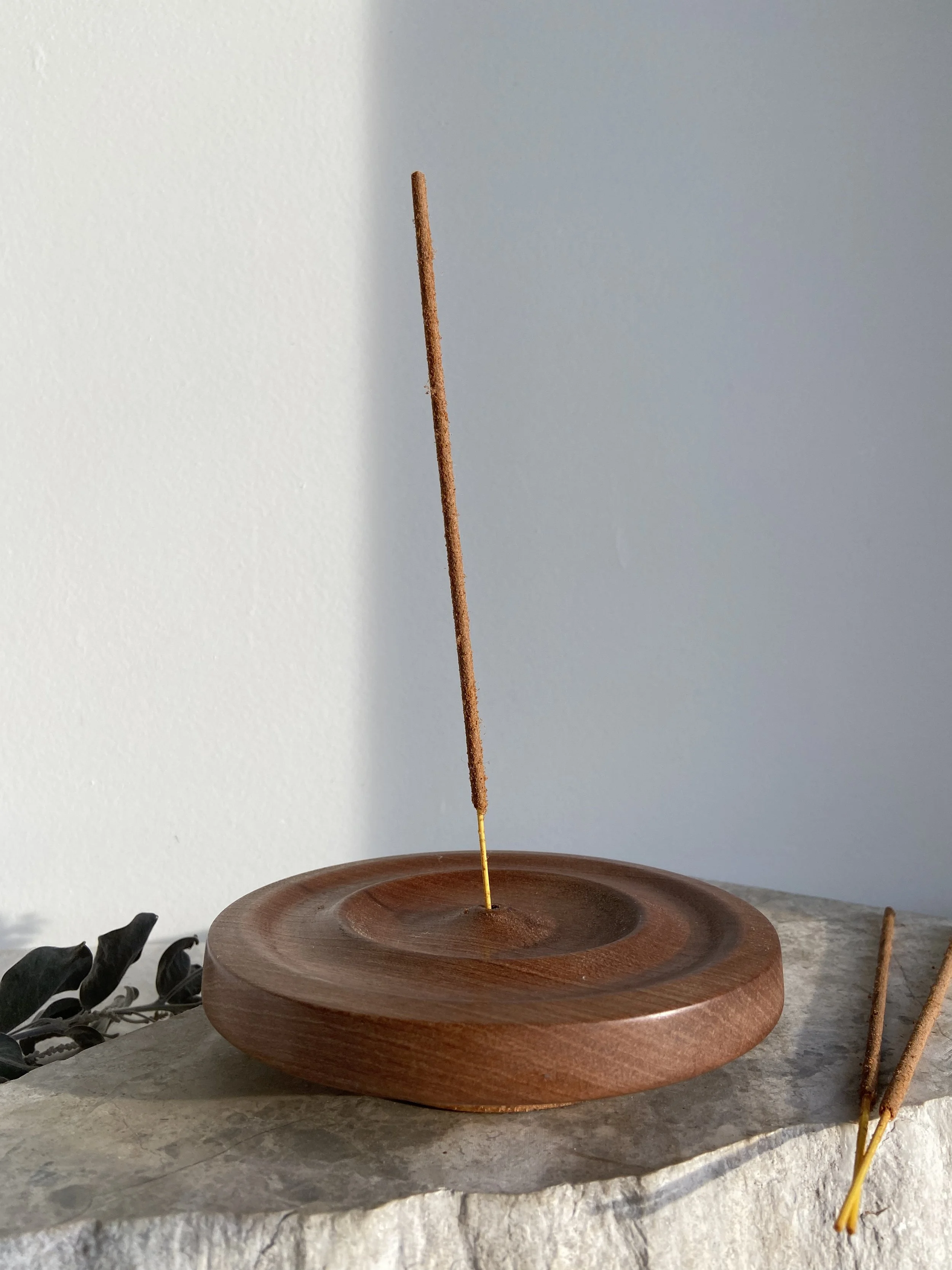 A wooden incense holder with one incense stick burning upright and two additional unlit incense sticks resting on a stone surface.