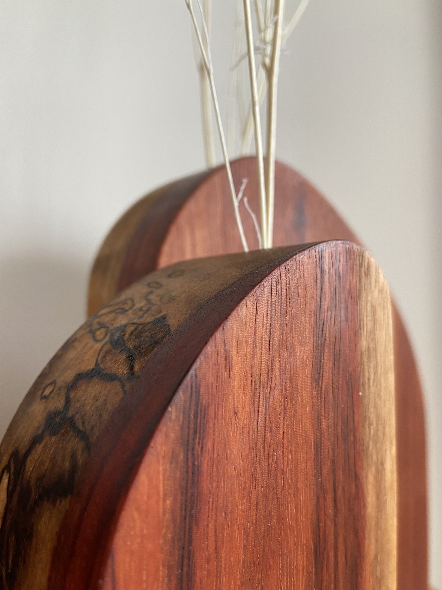 Close-up of wooden cutting boards in different shades, leaning against a light-colored wall with dried decorative grass attached.