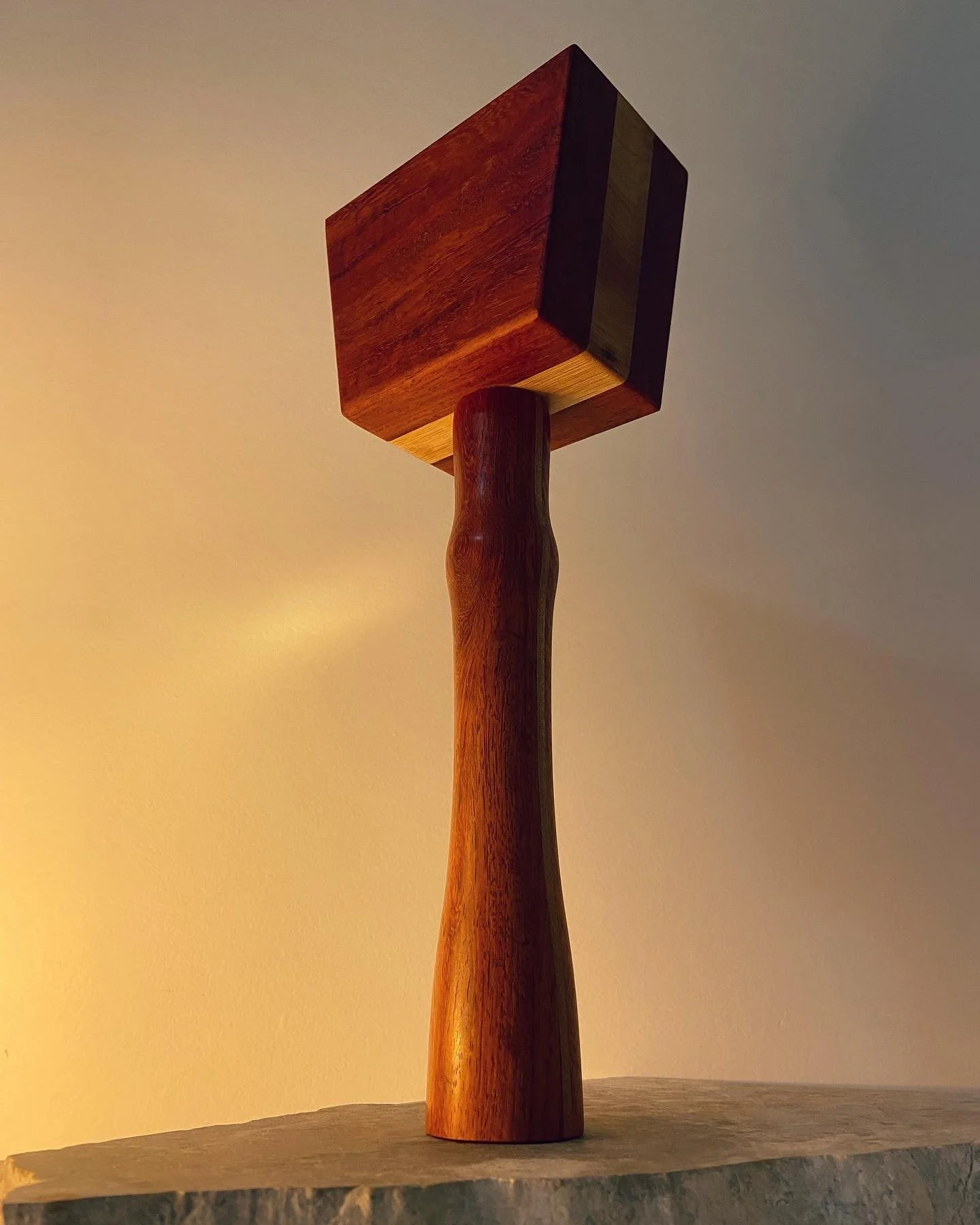 Wooden sculpture resembling a hammer with a thick handle and a square head, displayed on a piece of stone or concrete slab.