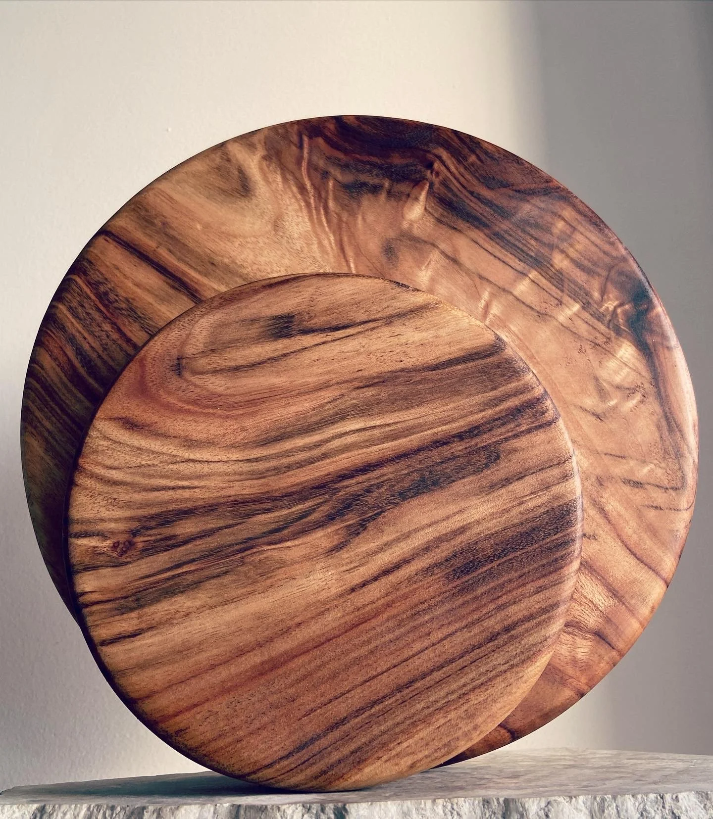Two round wooden bowls with natural wood grain patterns, one smaller and placed in front of a larger one, on a light-colored textured surface.