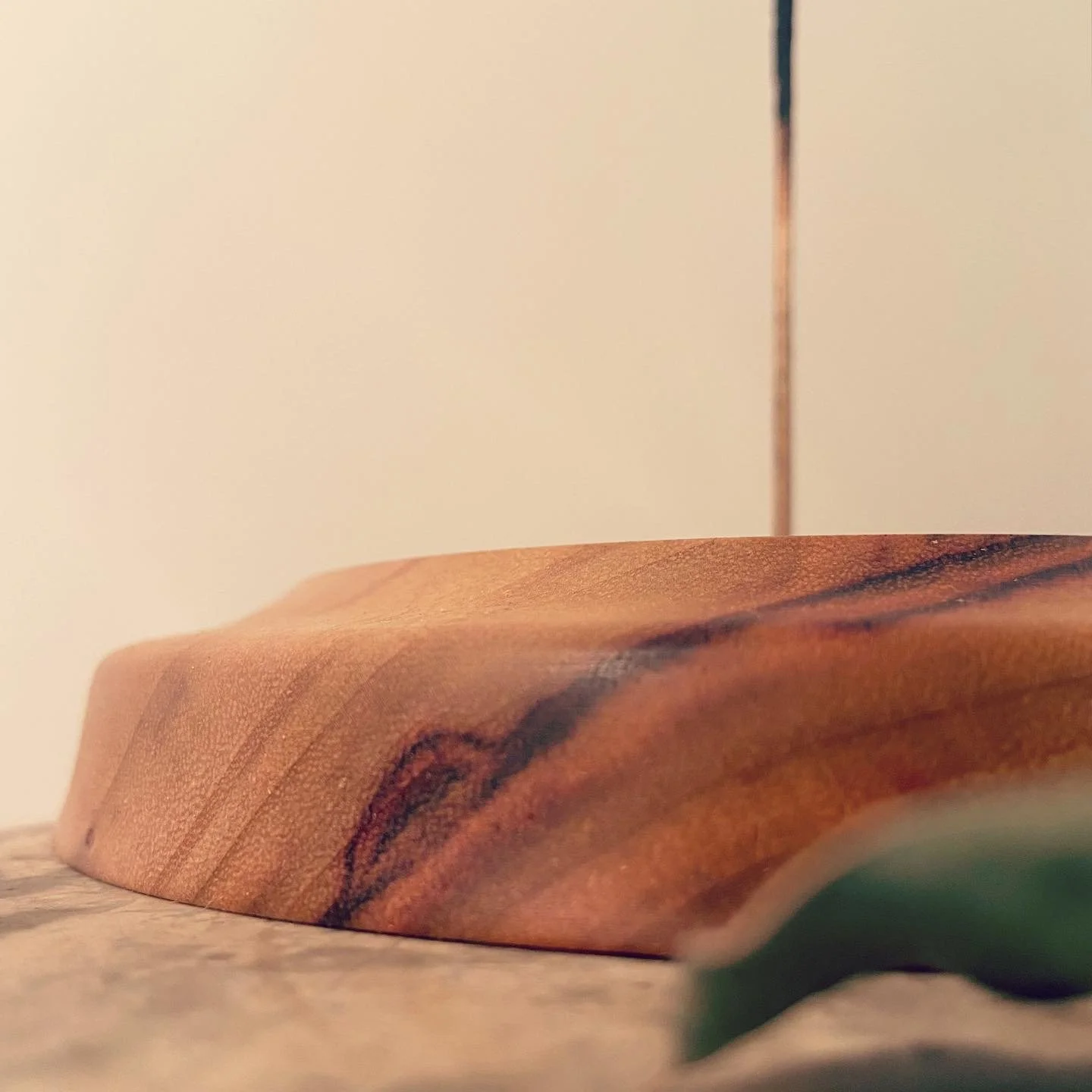 Close-up of a wooden cutting board with a smooth, polished surface and natural wood grain, resting on a textured surface against a plain background.