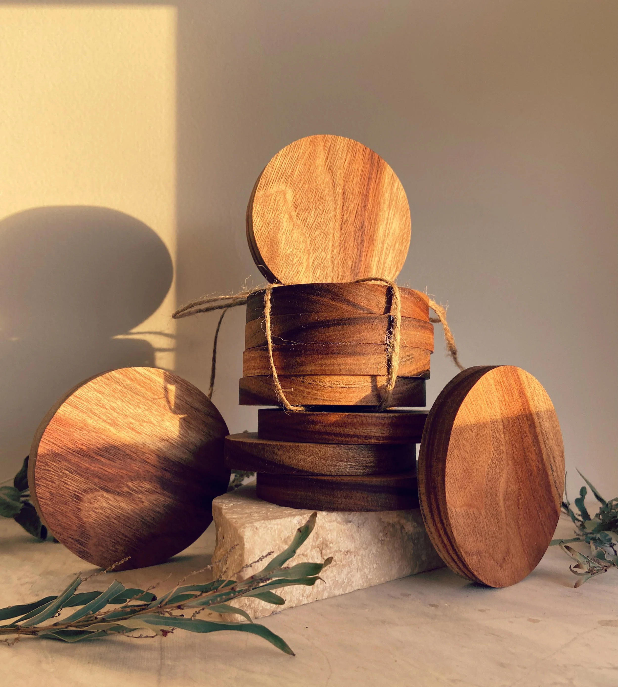 Stacked wooden coasters on a marble slab with greenery, lit by warm sunlight.