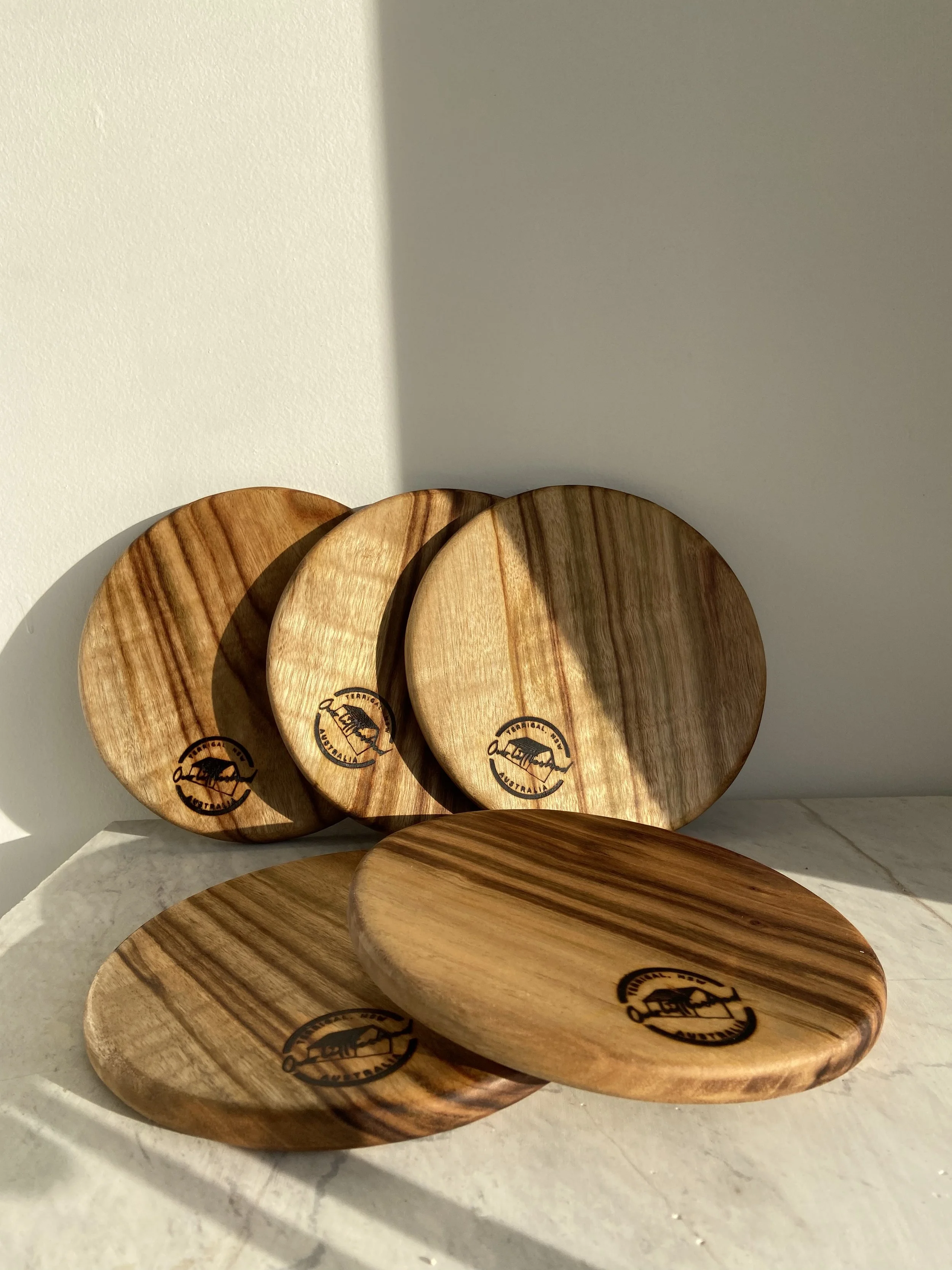 Five wooden plates with a logo, arranged on a white surface against a white wall.