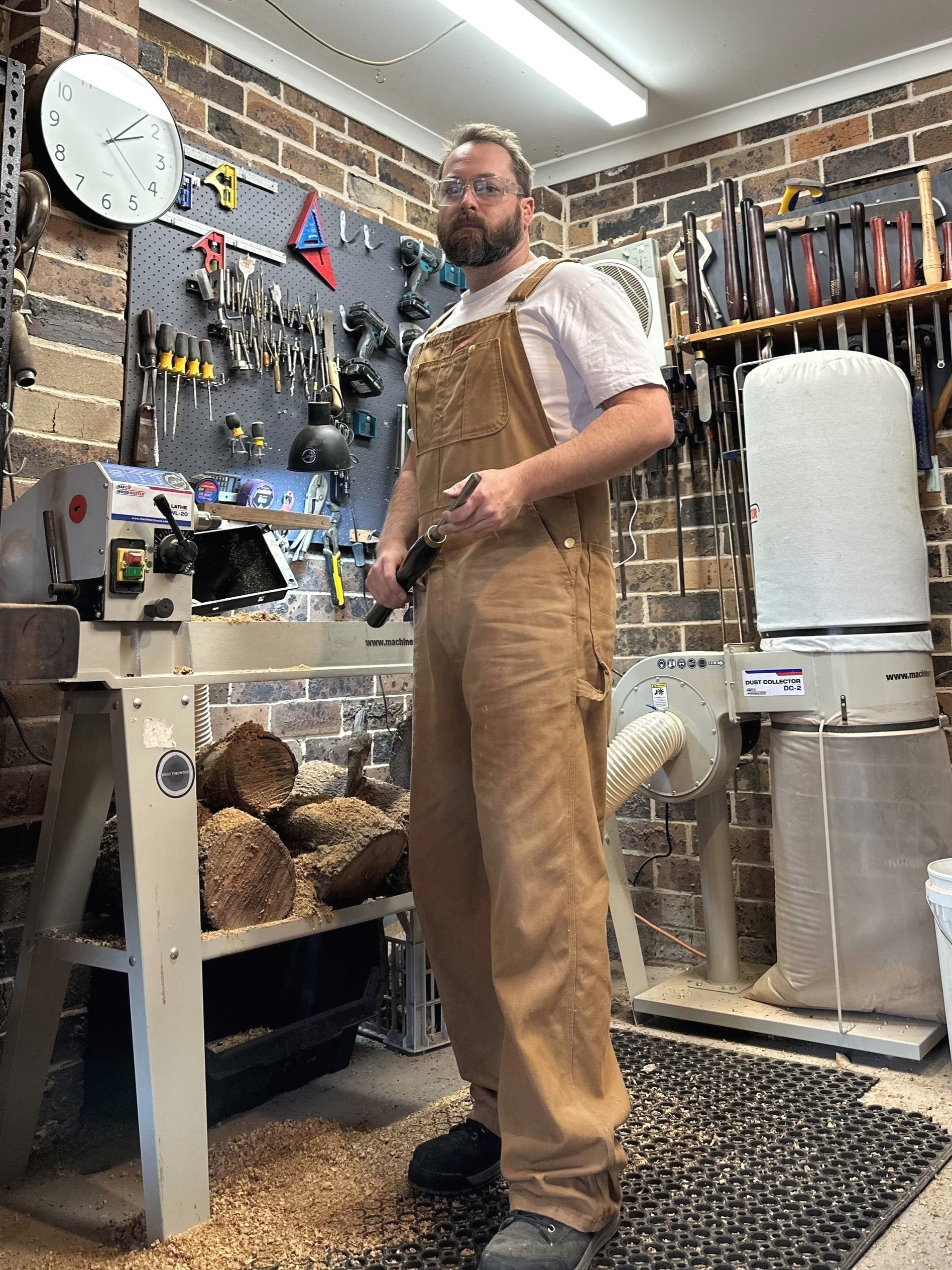 A man wearing safety glasses and tan overalls stands in a woodworking workshop, holding a hand tool, with a workbench, wood logs, and various tools on the wall behind him.