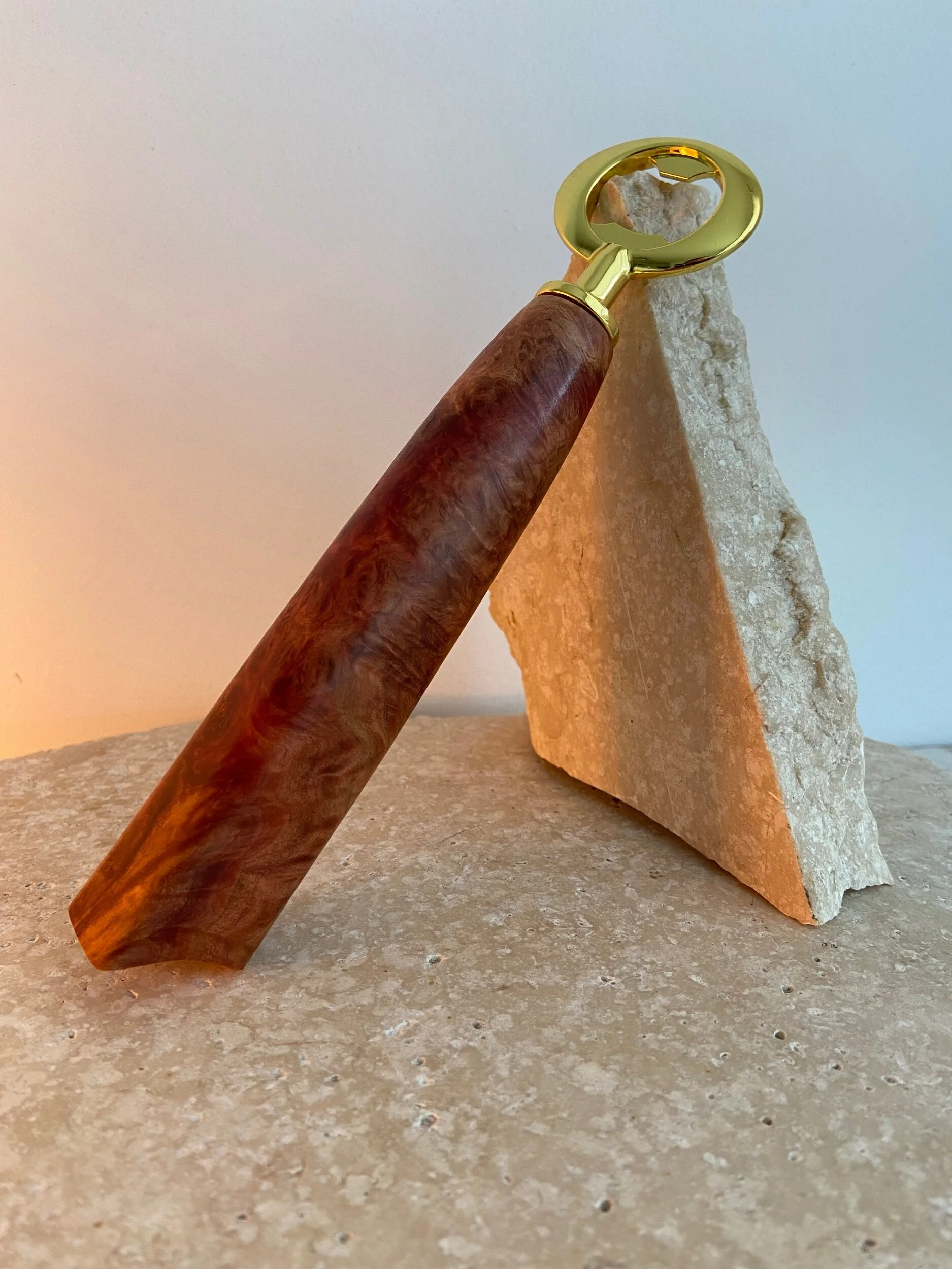 A bottle opener with a wooden handle and gold-colored metal head, leaning against a triangular piece of stone on a textured stone surface.