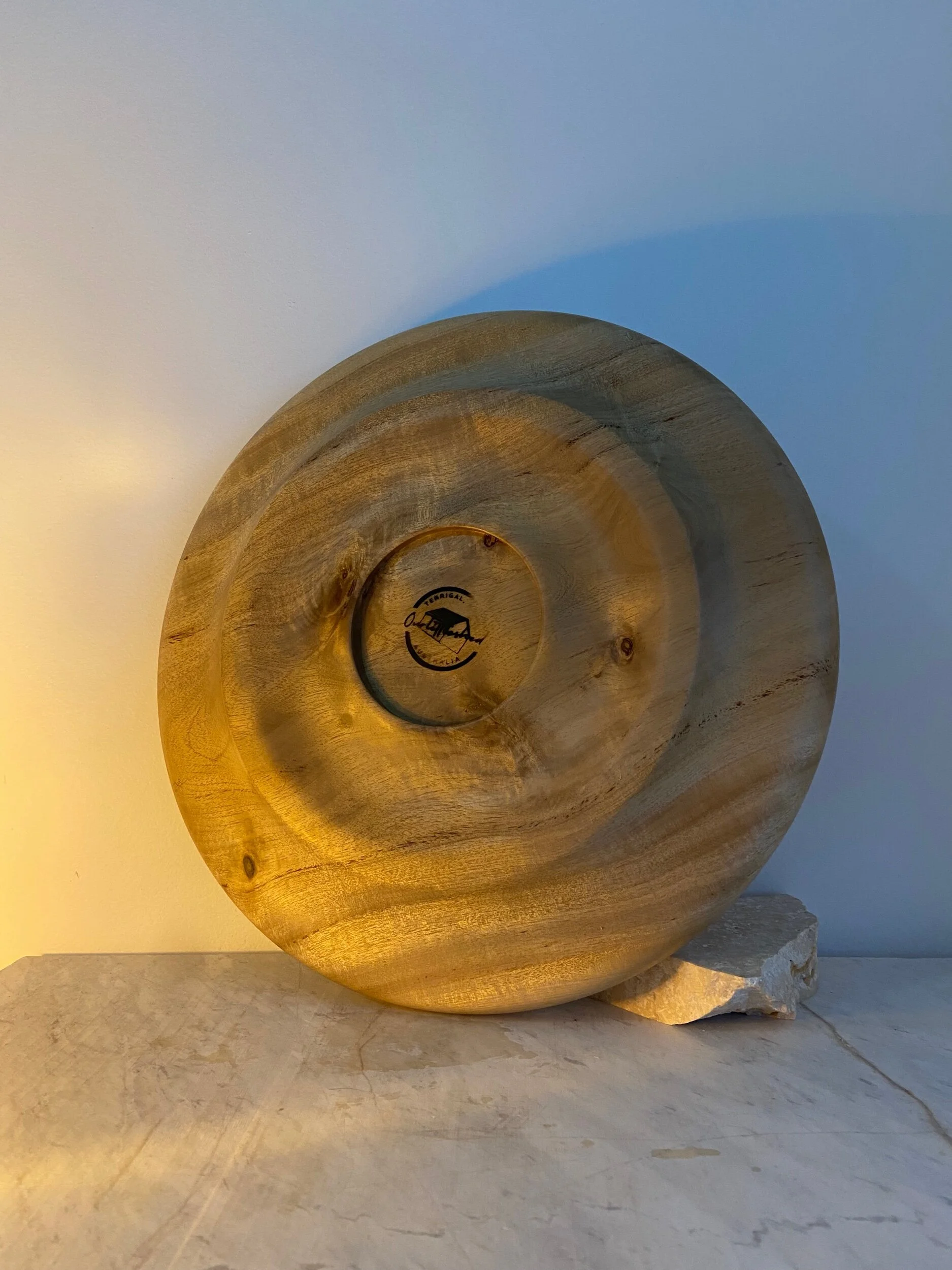A round wooden decorative piece with a carved spiral design, placed on a white surface against a white background, with a small rock at the bottom right