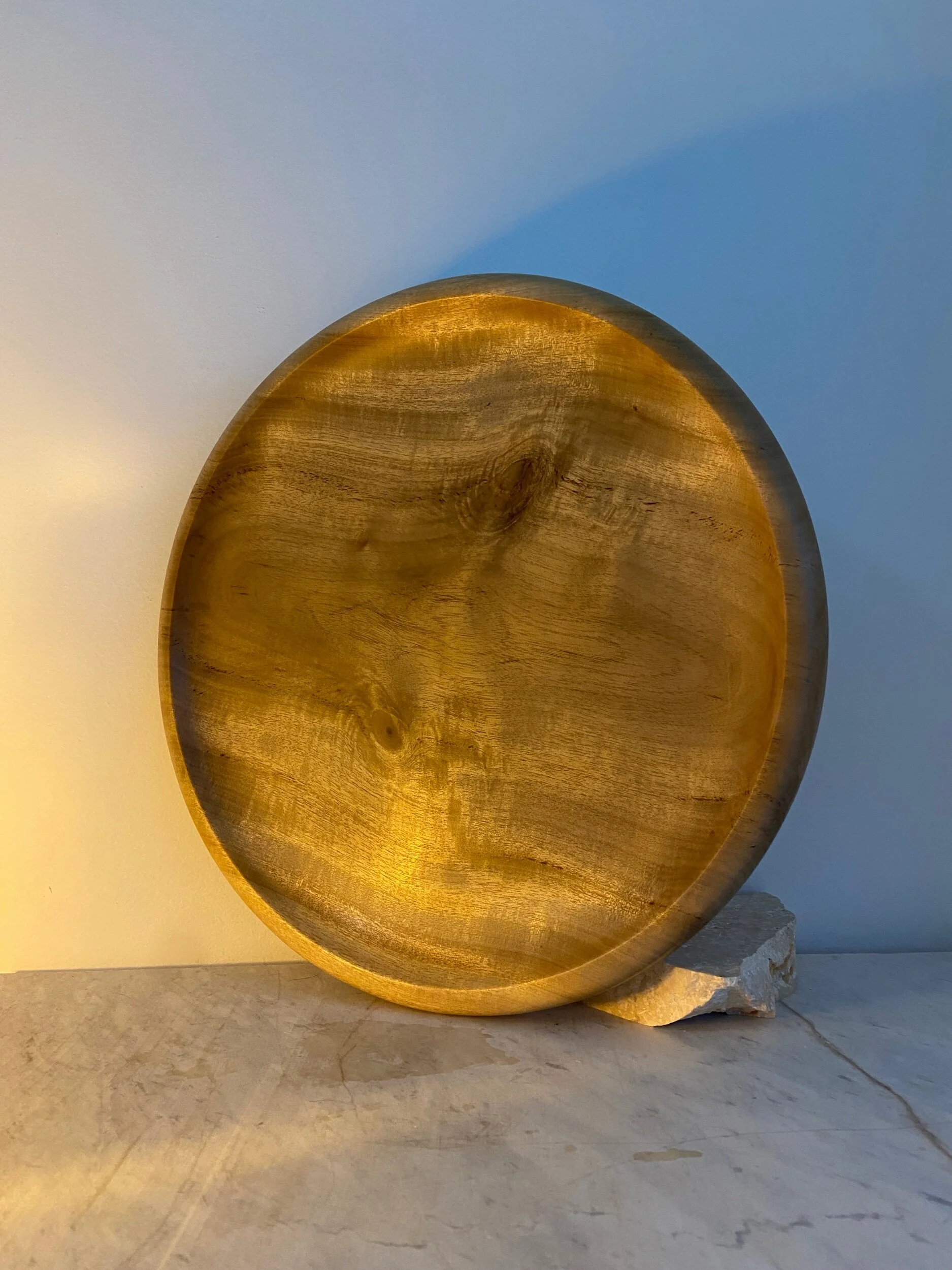 A round wooden bowl with a natural wood finish, placed on a light-colored surface against a plain wall.