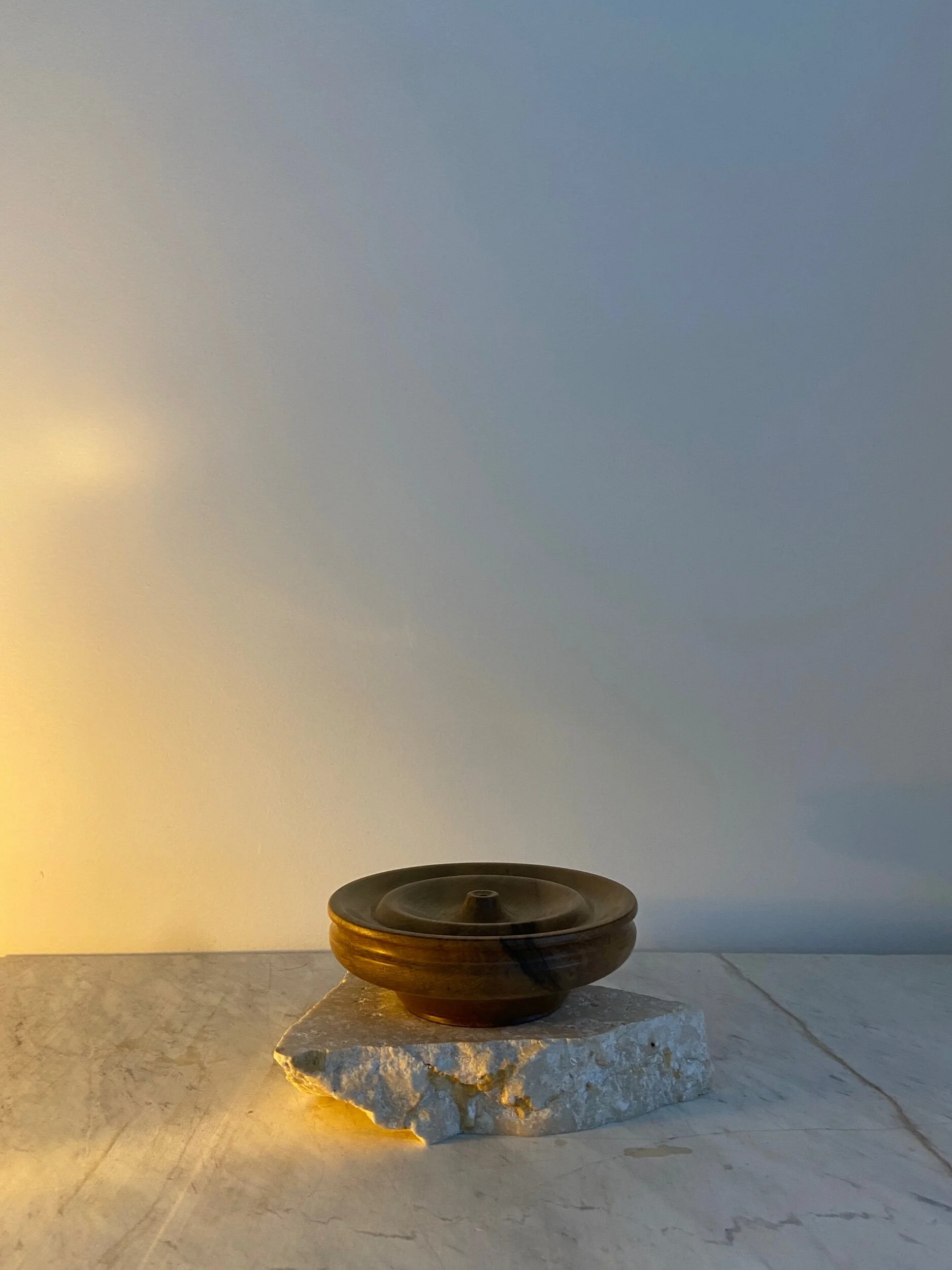 A dark brown ceramic bowl with a lid, placed on a rough stone slab, on a marble surface with a plain, light-colored background.