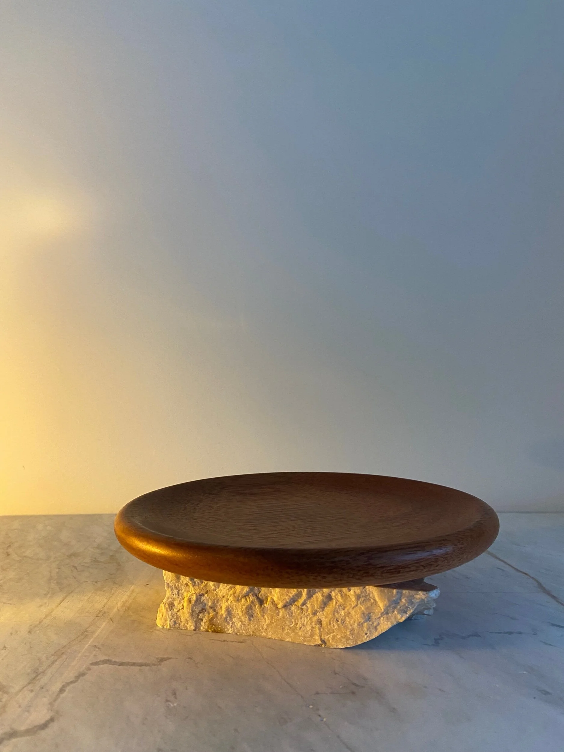 A round wooden plate resting on a rough, white stone slab, with soft lighting creating a warm shadow on a marble surface.