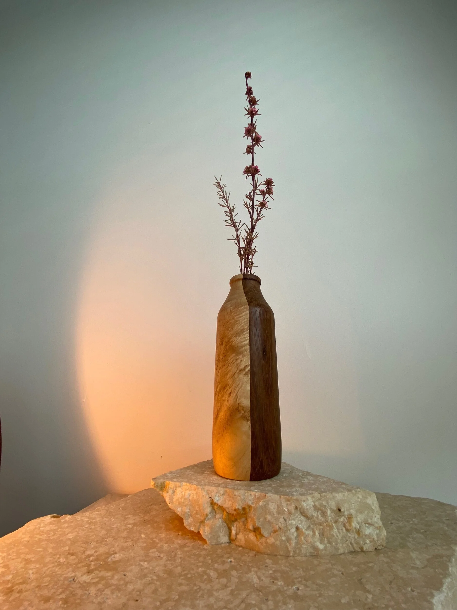 Wooden vase with a slender, neutral-colored branch with small flowers, placed on a rough stone surface against a light wall.