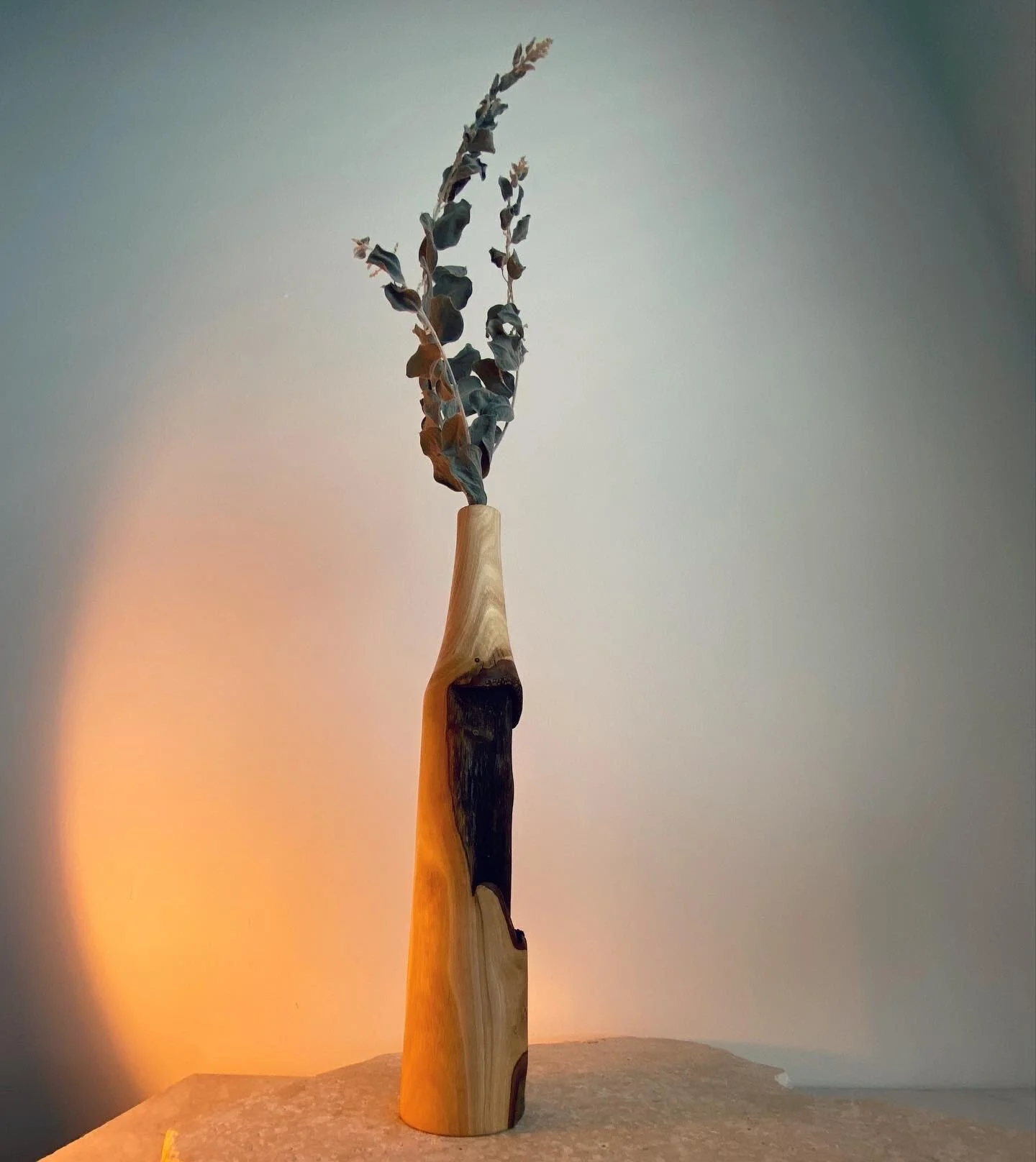 A tall wooden vase with a natural finish holding a dried plant with withered leaves, placed on a textured surface against a plain background.