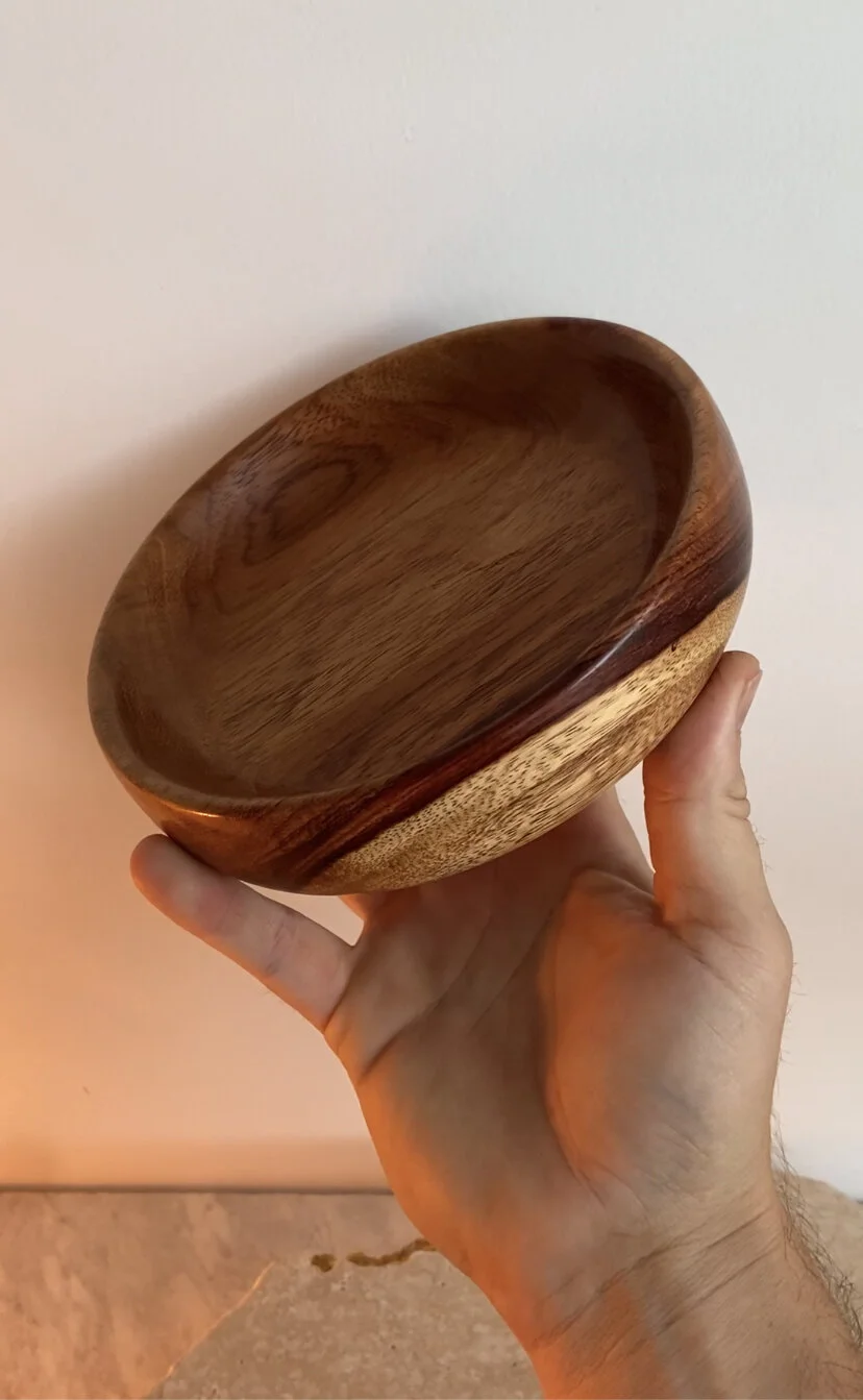 A hand holding a round wooden bowl with a natural wood grain pattern against a plain wall background.