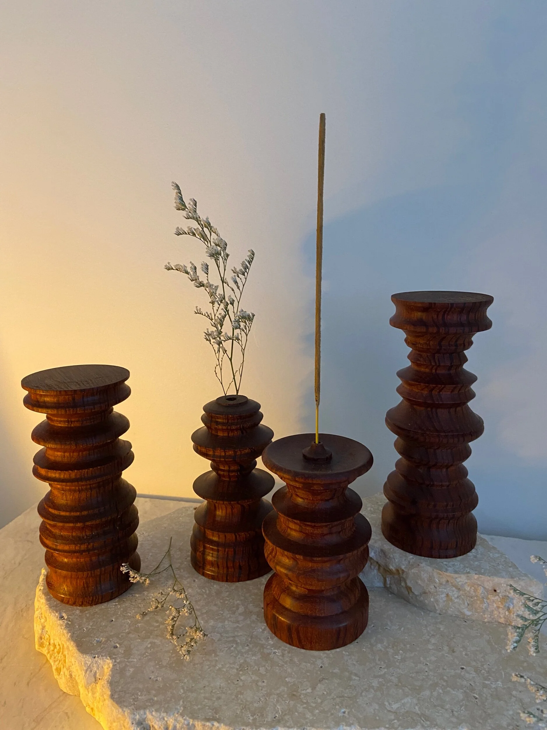 Five distinctive wooden candle holders of varying heights, with one holding a dried flower and an incense stick, placed on a rough stone surface against a wall with a gradient from light to darker blue.