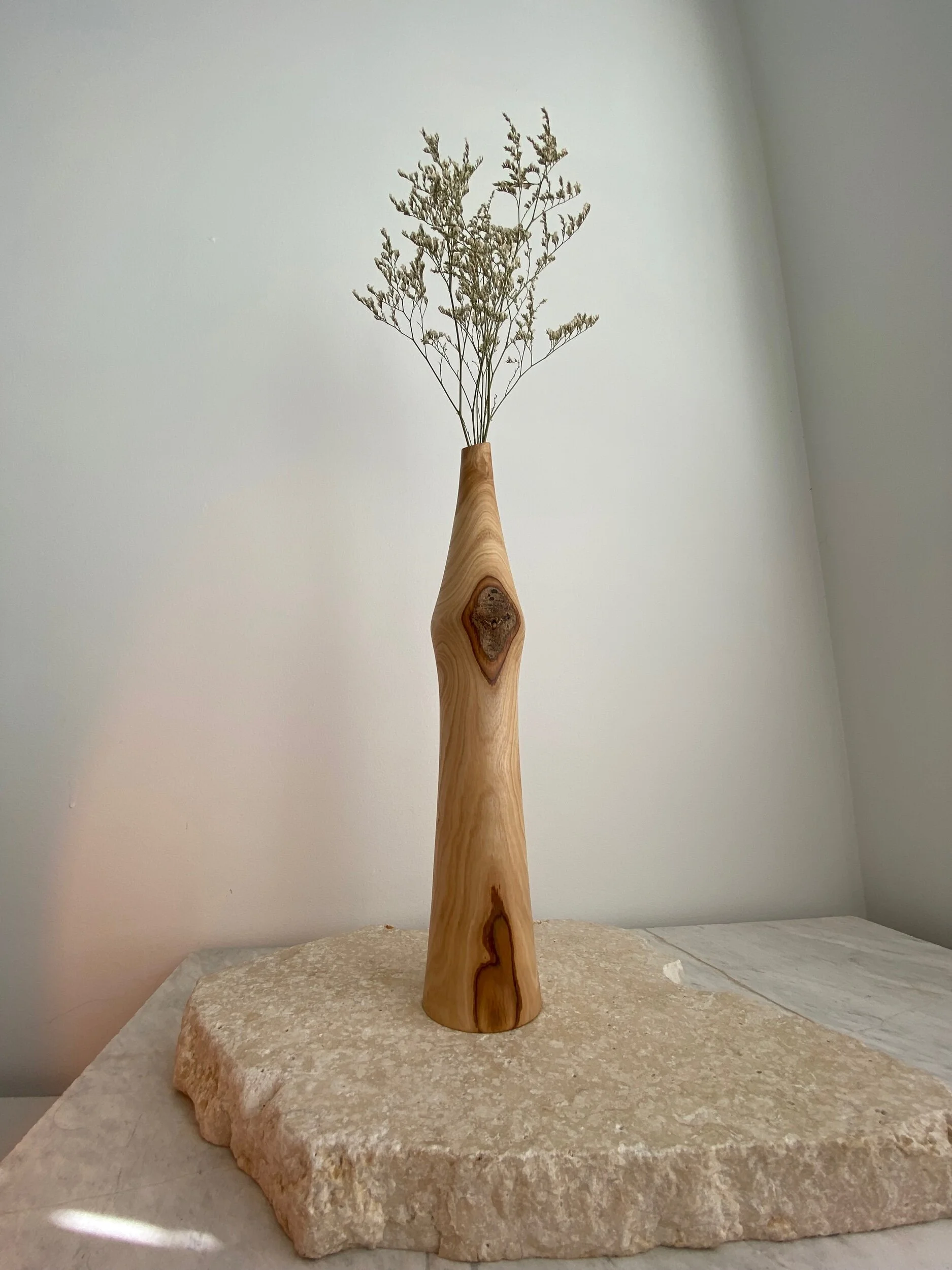 Wooden vase with a narrow neck holding dried white flowers, placed on a rough stone slab against a white wall.