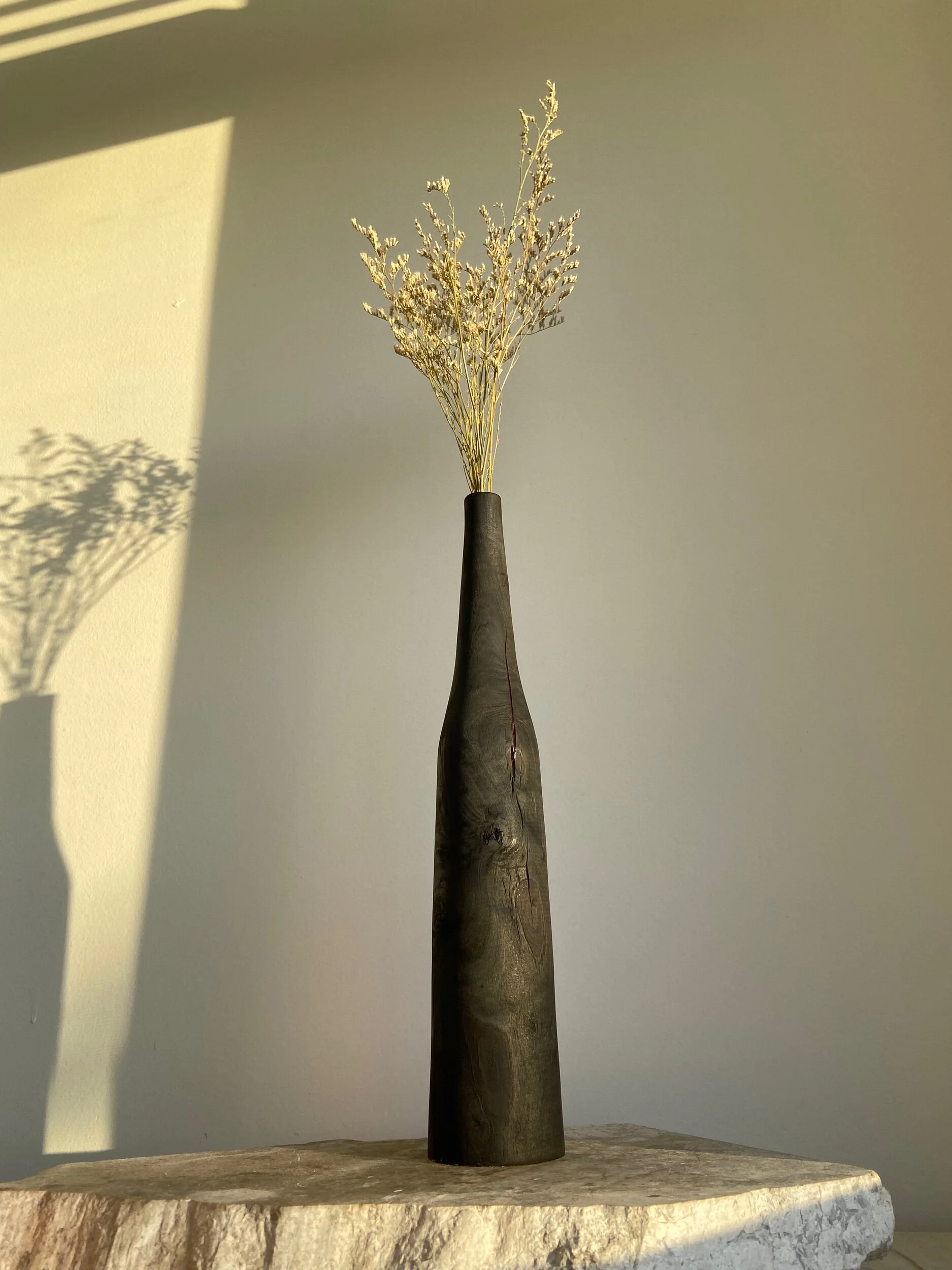 A tall, dark wooden vase with a narrow neck, holding a bunch of dried, pale-colored sprigs, placed on a textured stone surface with soft shadows on a plain wall background.