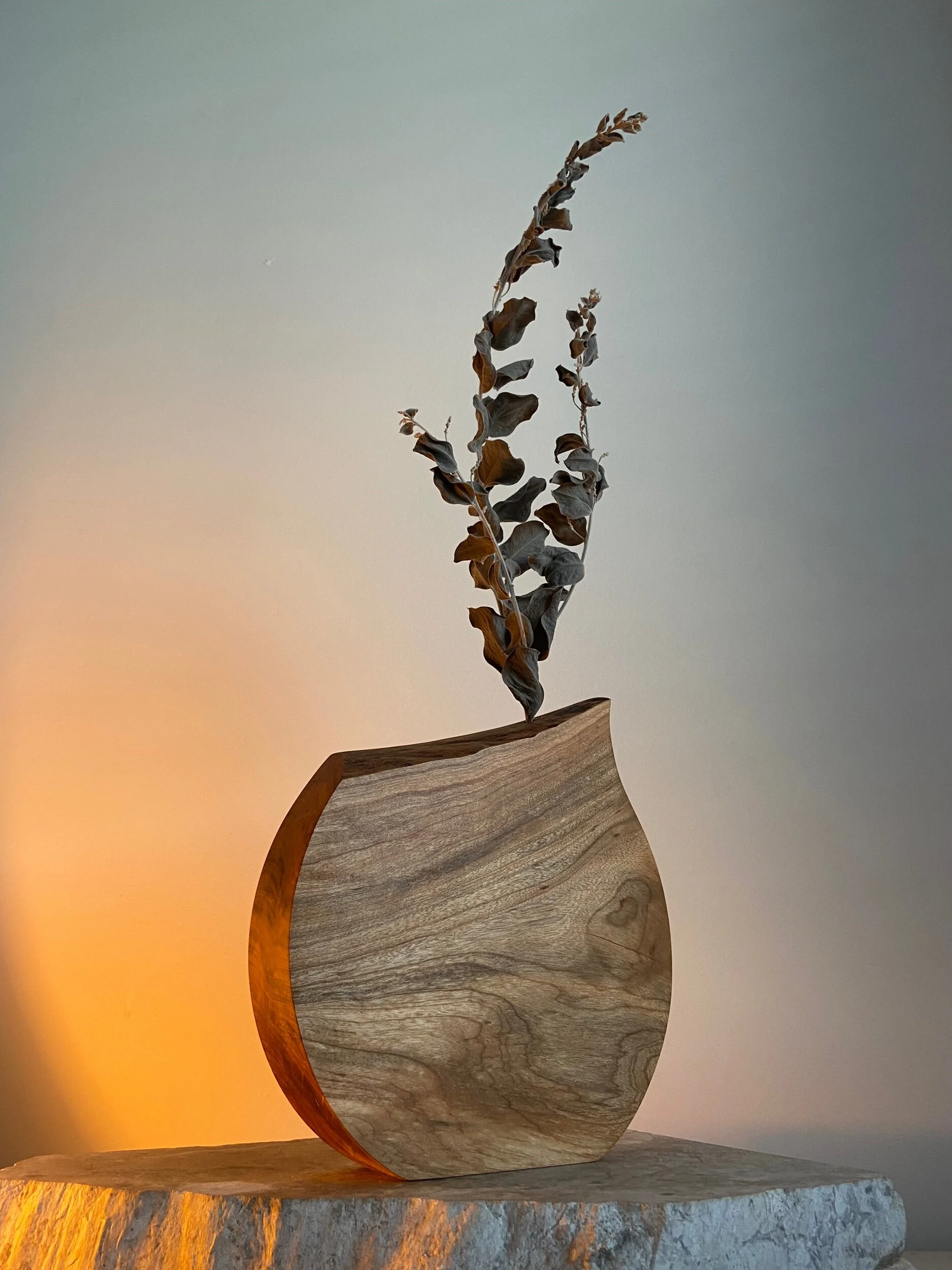A wooden vase with a curved shape holding a dried brown and gray branch, placed on a textured stone surface against a neutral background with soft lighting.