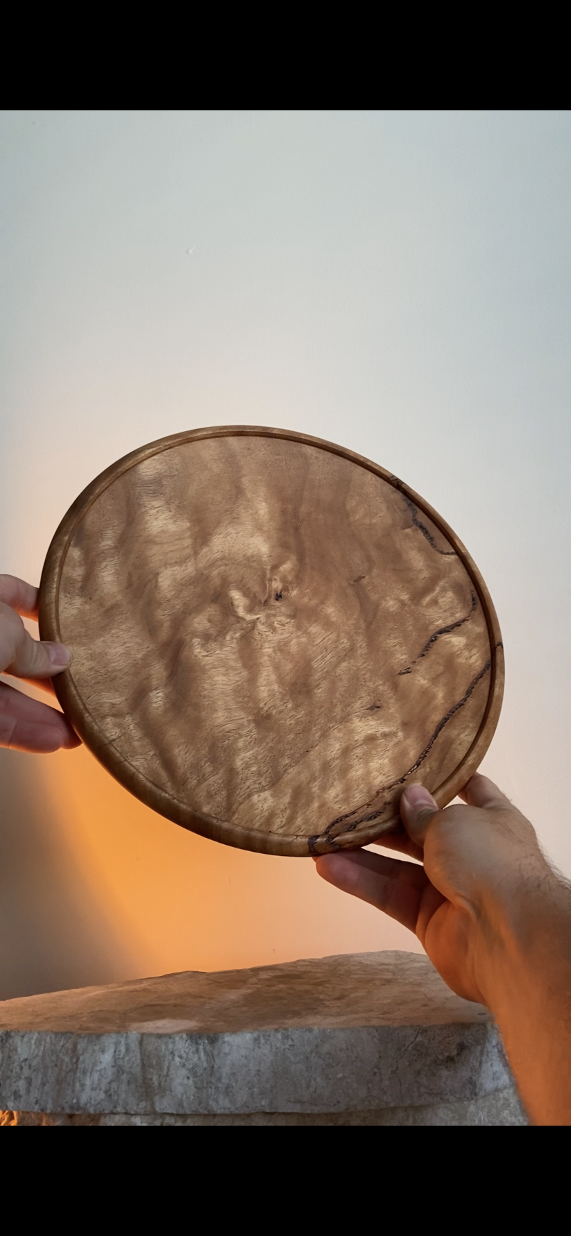 A person holding a round wooden plate with a natural wood grain pattern and a dark crack along one side, against a plain background.