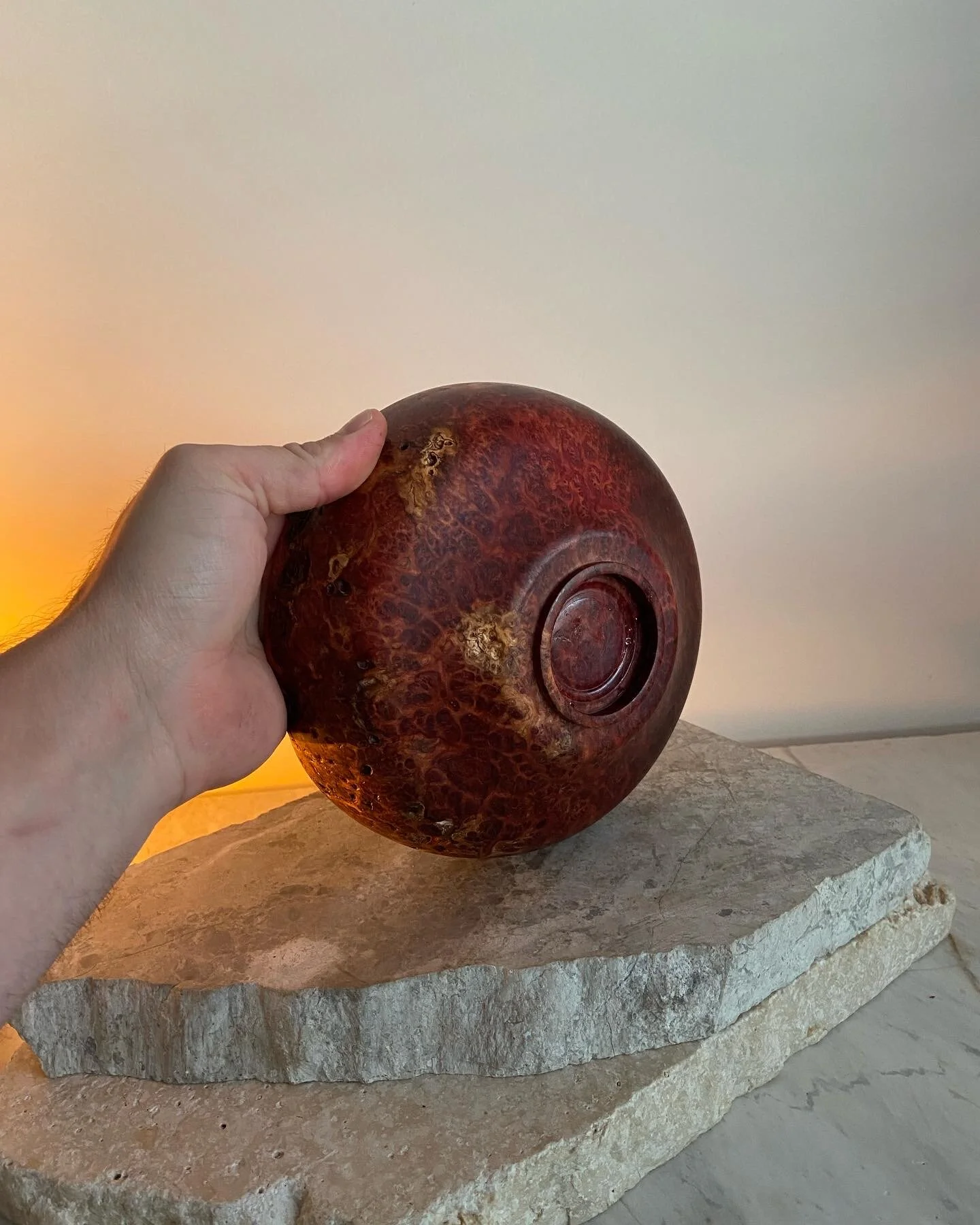 A hand holding a round, polished, dark reddish-brown decorative object with a textured surface, placed on a stone slab against a plain background.