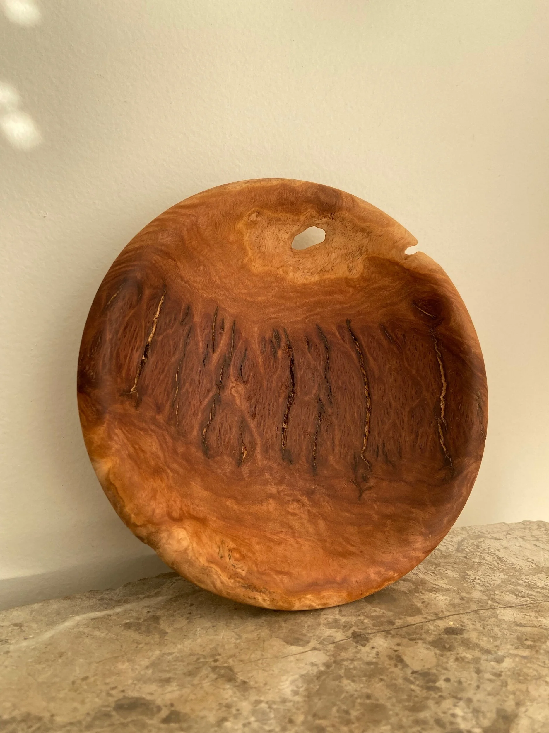 A round wooden bowl with a hole near the edge, sitting on a beige stone surface against a light-colored wall.